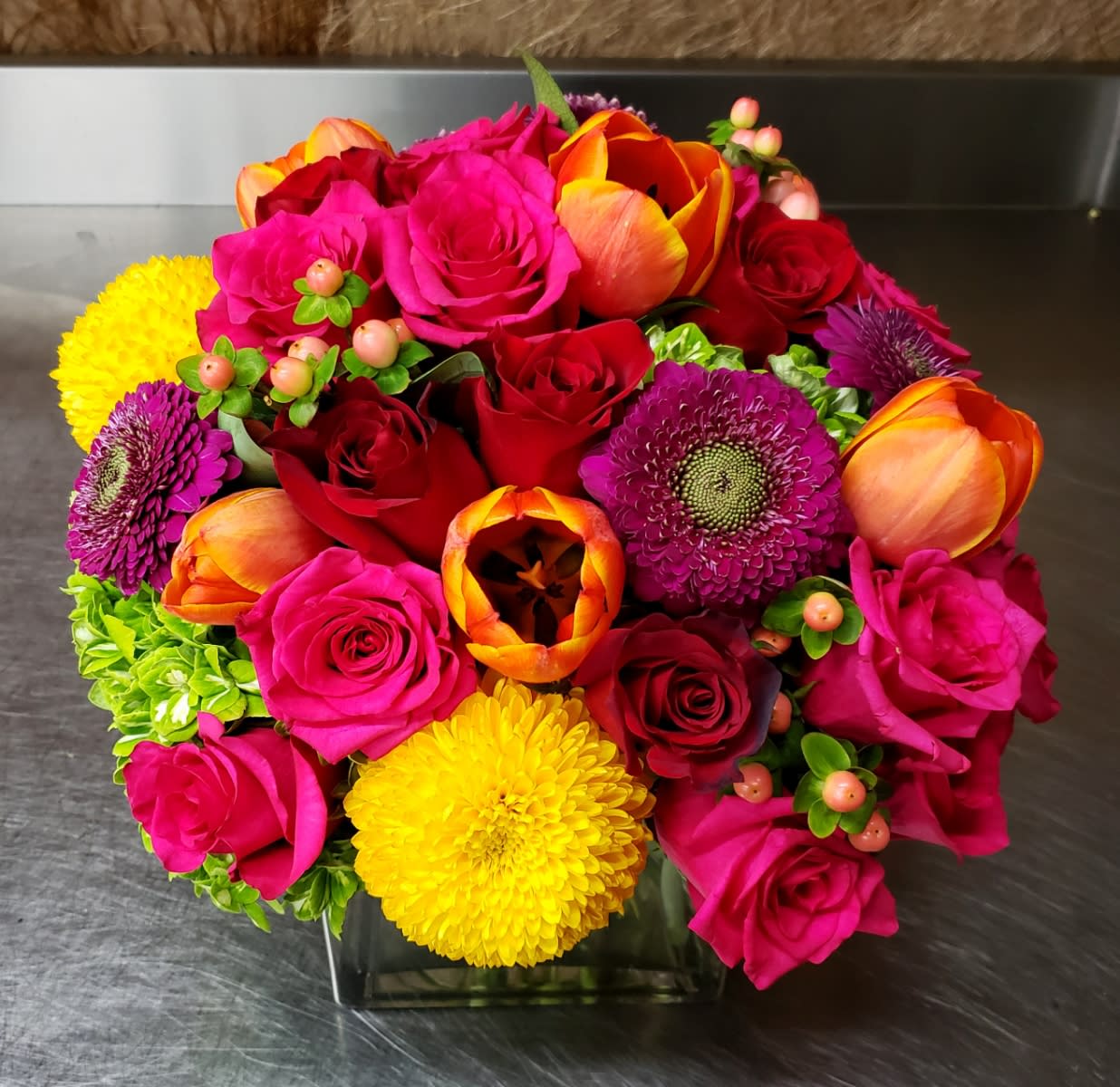 Bright mixed bouquet of roses, tulips, and mums in a square glass vase