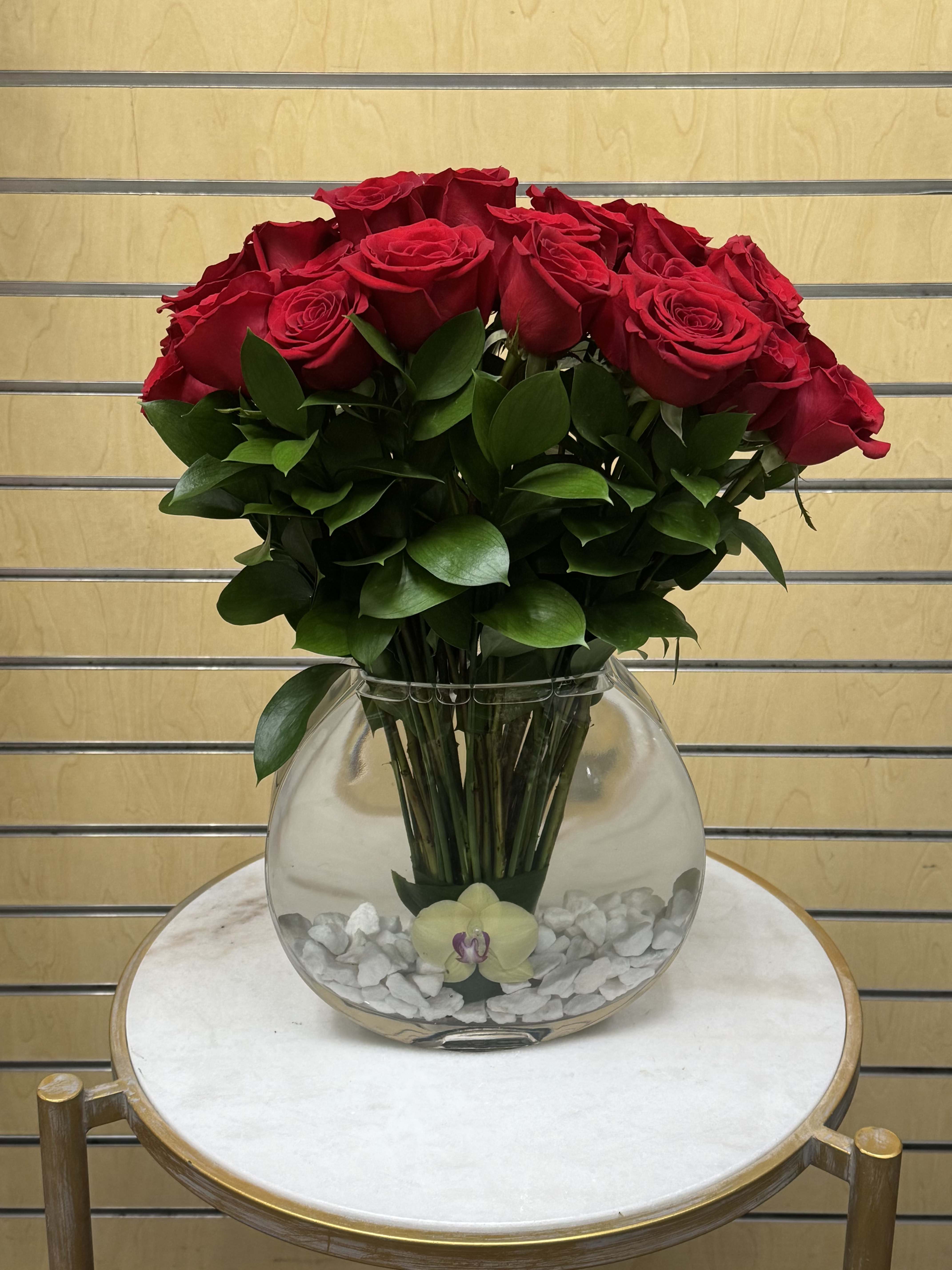 Red roses arranged in a clear round glass vase with white stones.