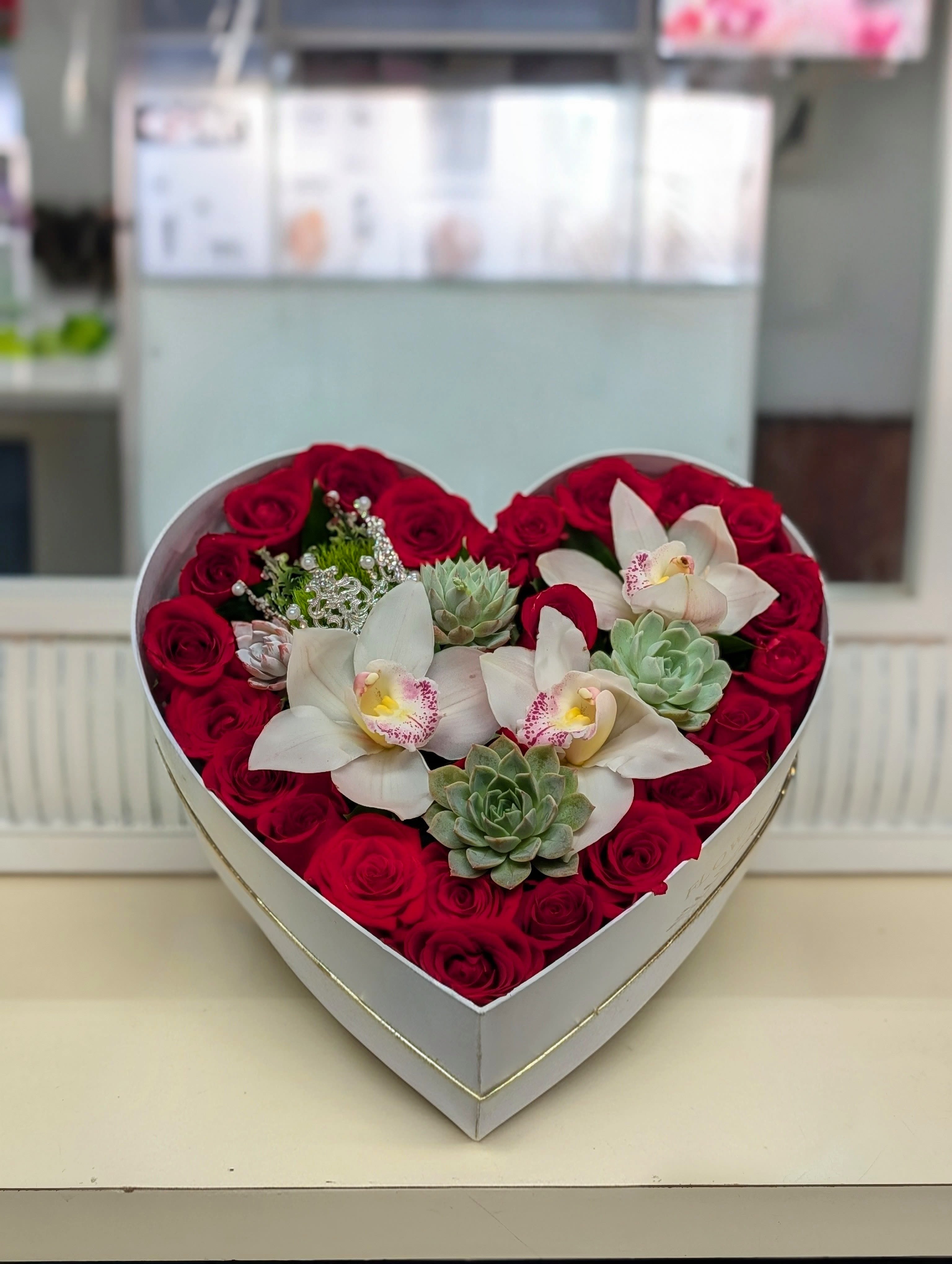 Heart-shaped box with red roses, white orchids, and succulents arranged tightly together