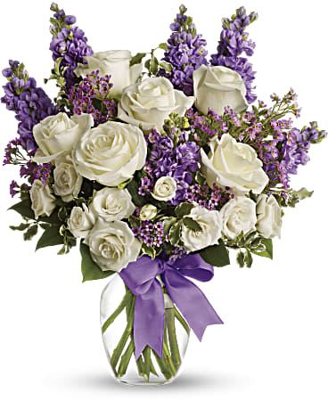 White roses and purple flowers arranged in a glass vase with a purple ribbon