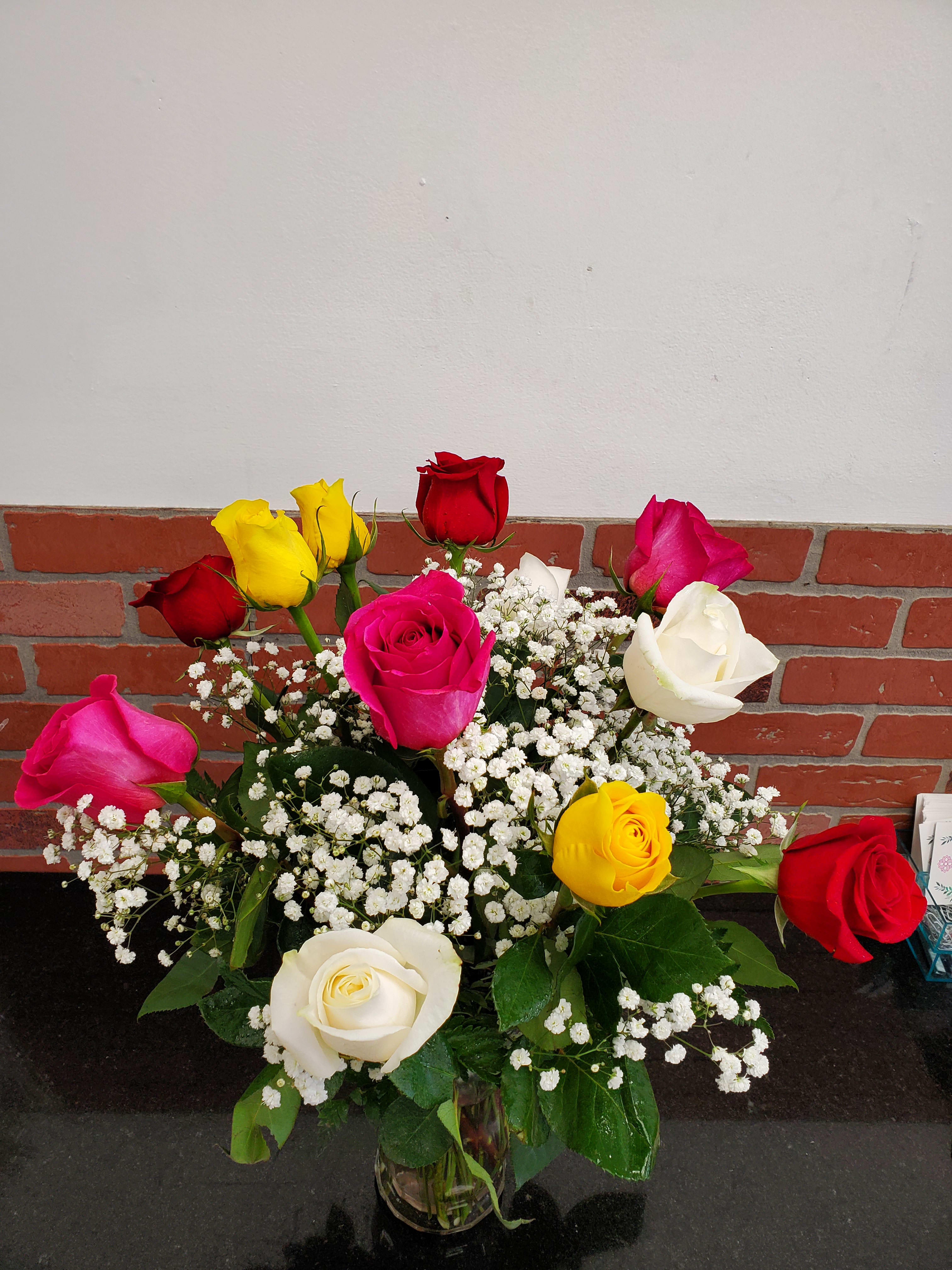 Mixed red, pink, yellow, and white roses with baby's breath in a clear glass vase on a dark countertop
