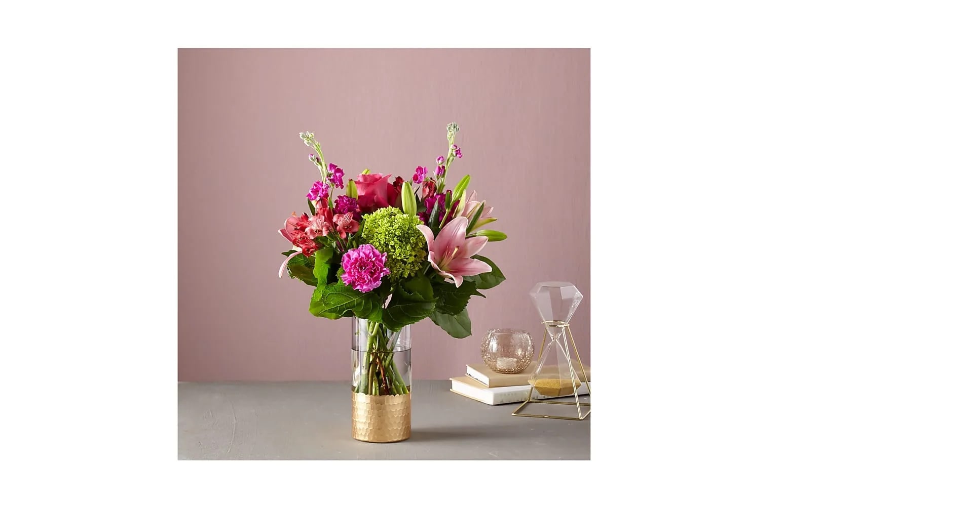 Mixed pink lilies, roses, carnations, and green hydrangea in a clear vase with gold base on a tabletop