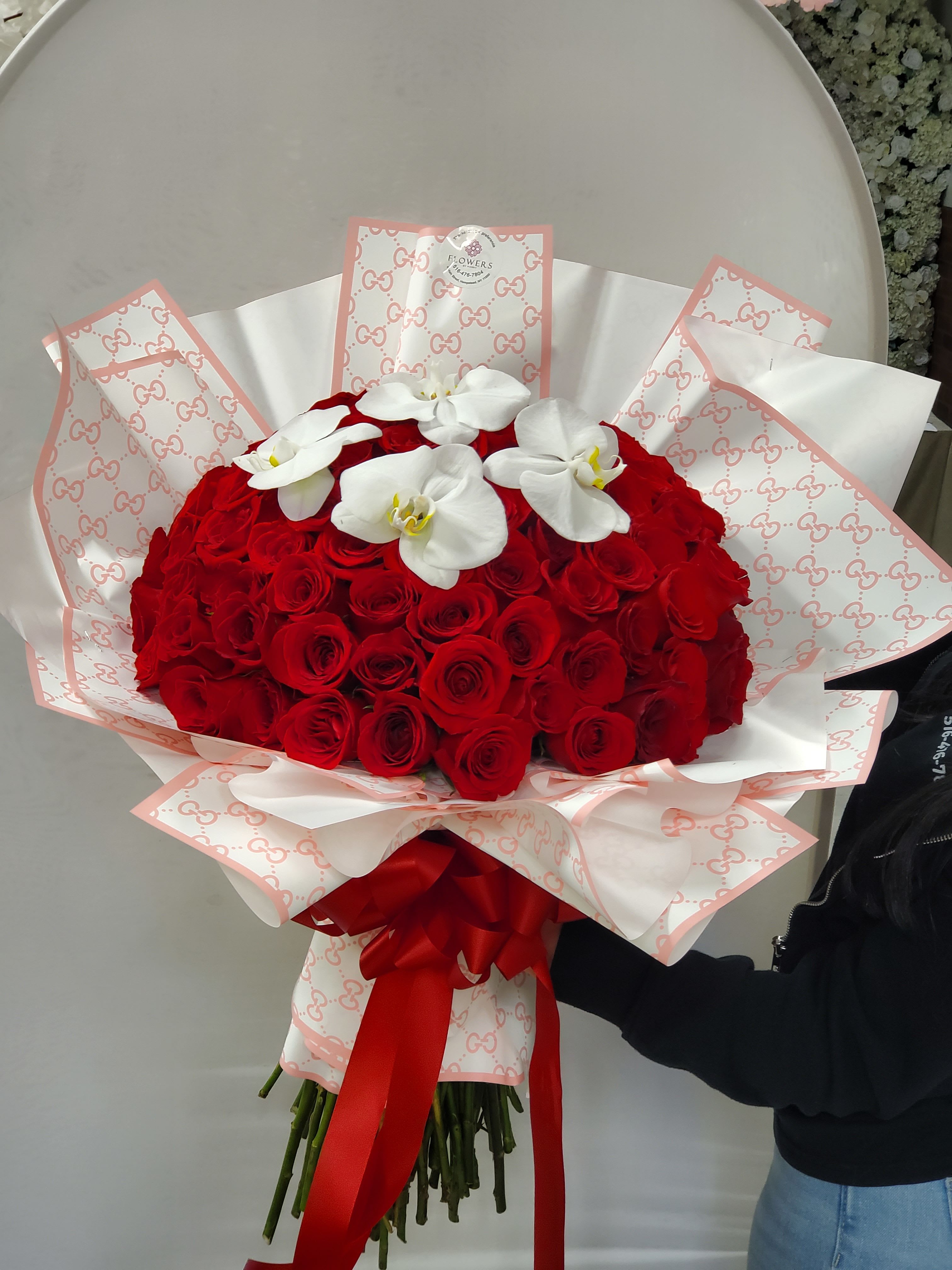 Large hand-tied bouquet of red roses with white orchids, wrapped in patterned paper and tied with a red bow.