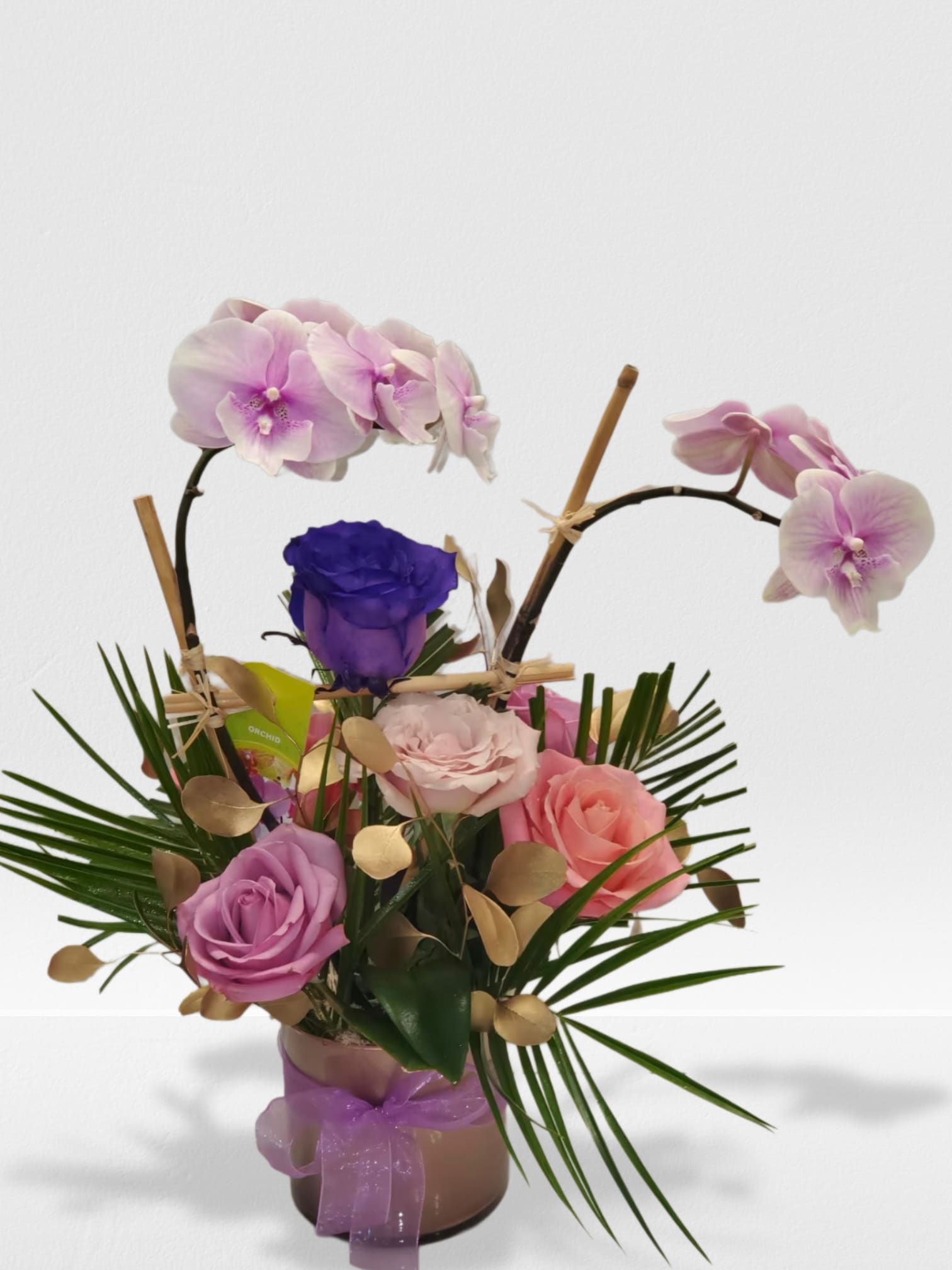 Arrangement of pink orchids and mixed roses in a cylinder vase with palm accents and a purple ribbon