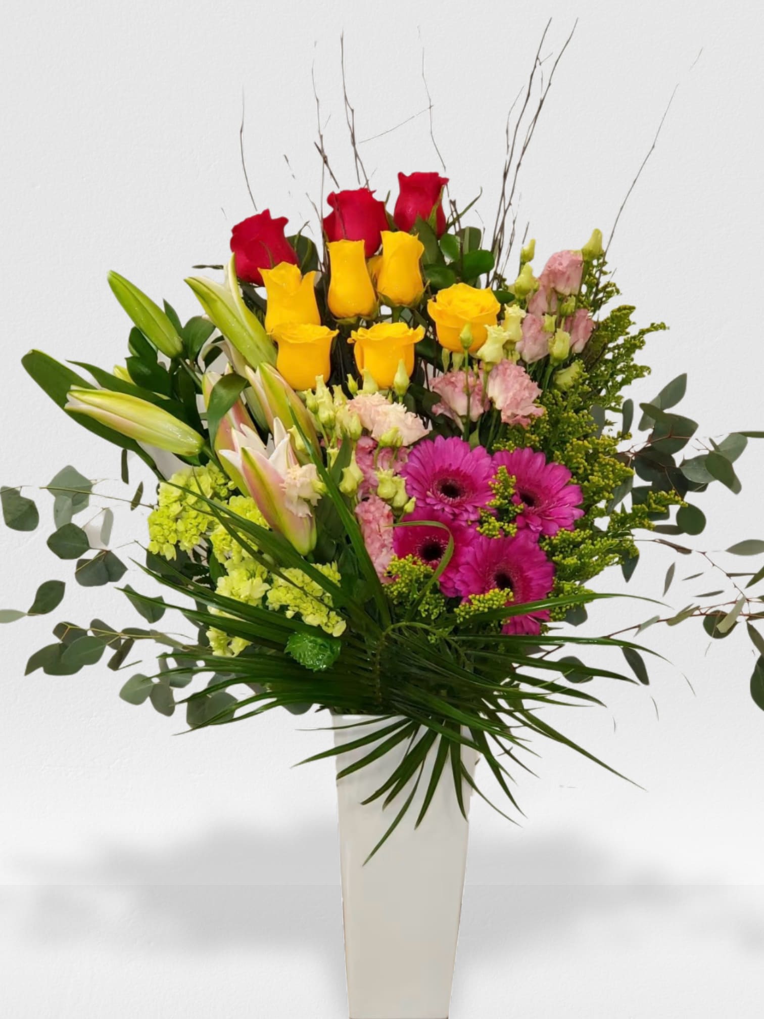 Tall mixed bouquet of red and yellow roses, pink lilies and gerberas in a white vase