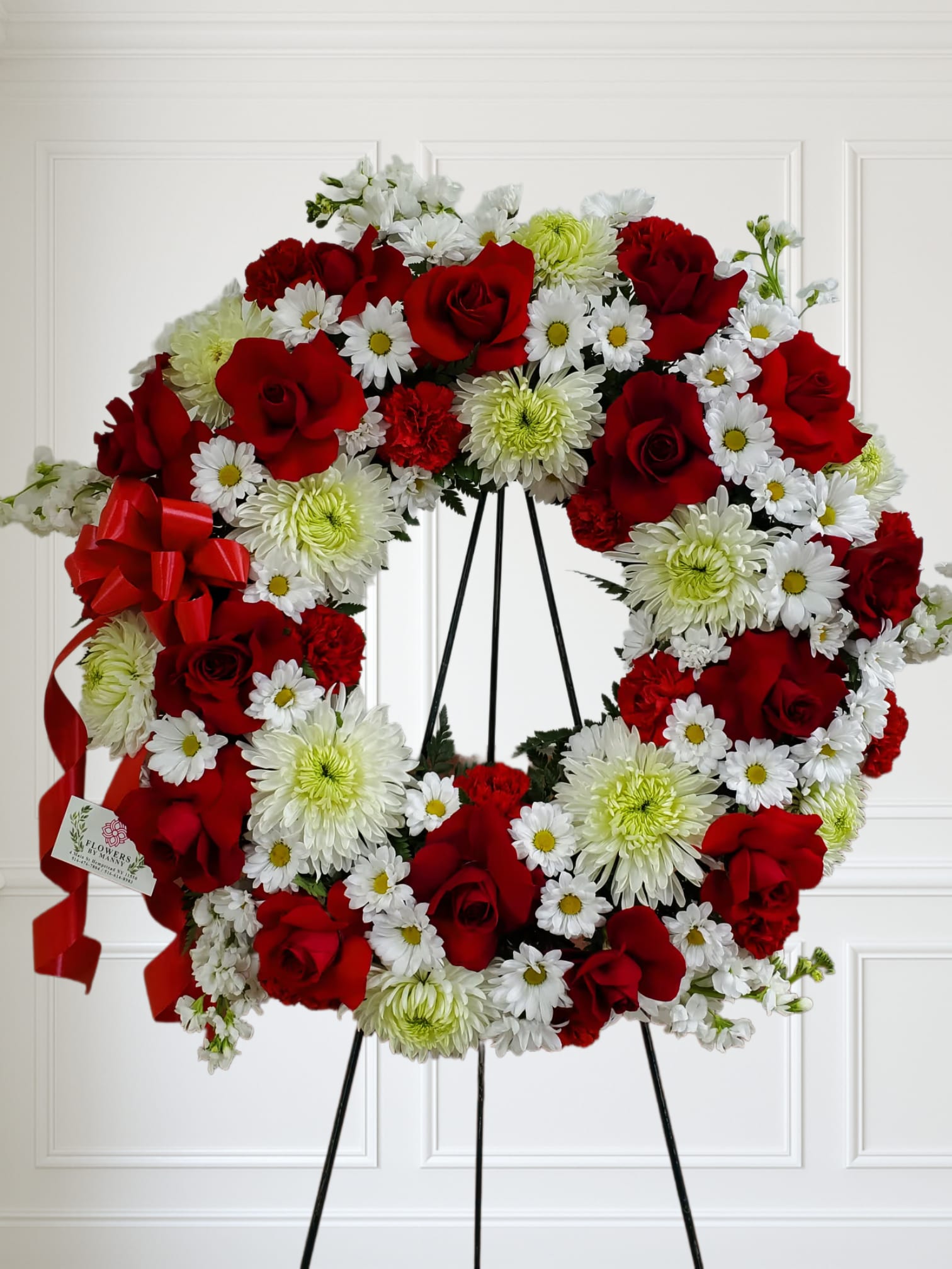 Standing wreath of red roses, white daisies, and green-tinted mums on a black easel with red ribbon bow