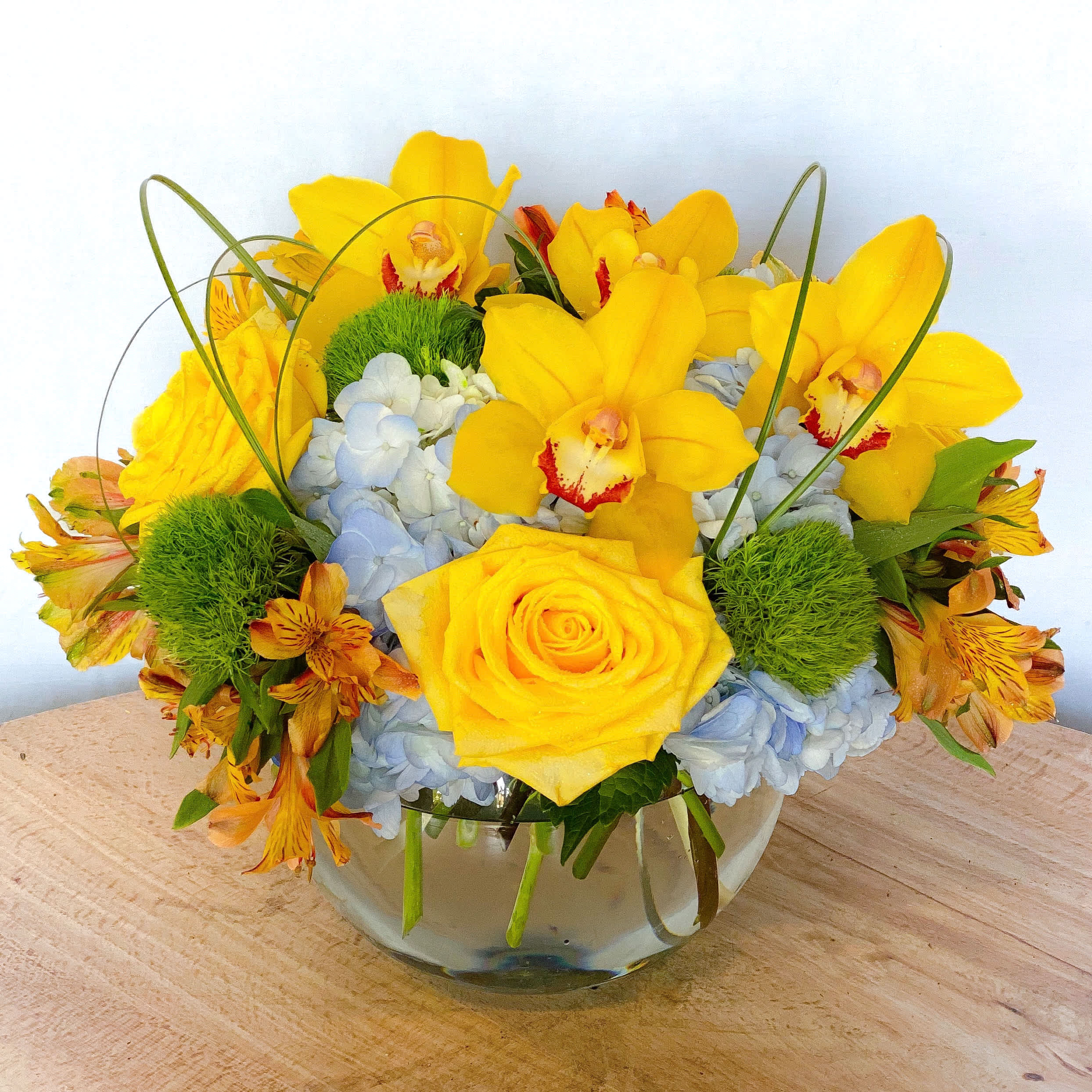 Yellow orchids and roses arranged in a glass bowl with blue hydrangeas