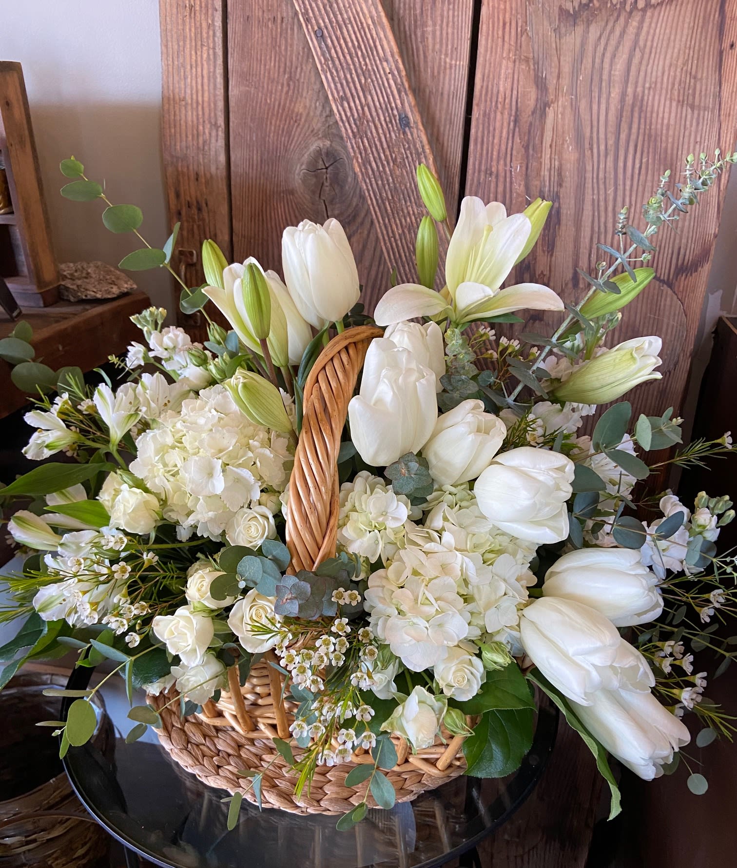 White tulips, lilies, and hydrangeas arranged in a woven basket against a rustic wooden backdrop