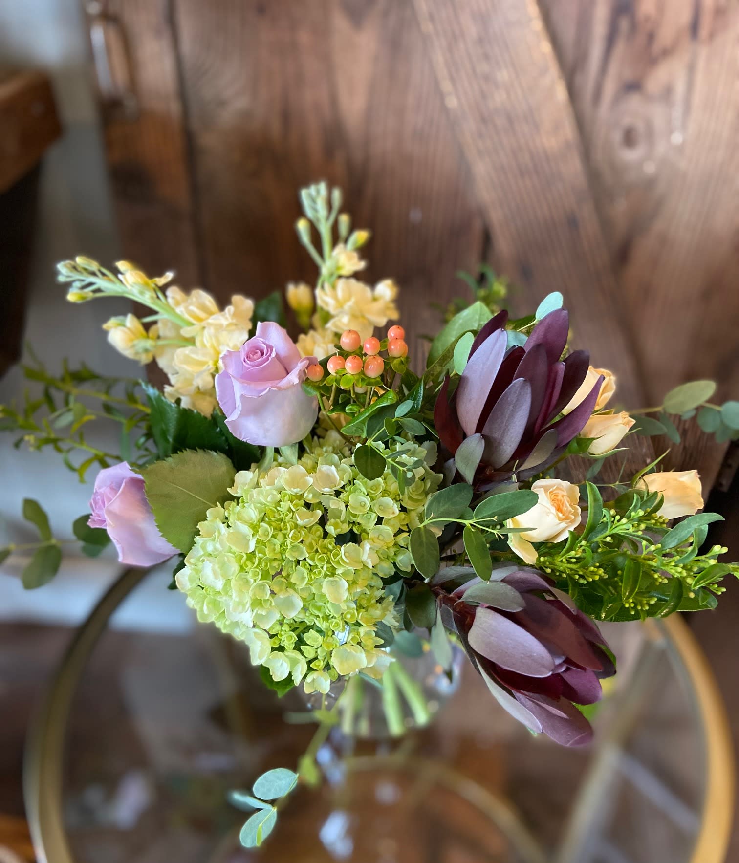 Arrangement with green hydrangea, lavender roses, cream blooms and dark burgundy protea in a glass vase
