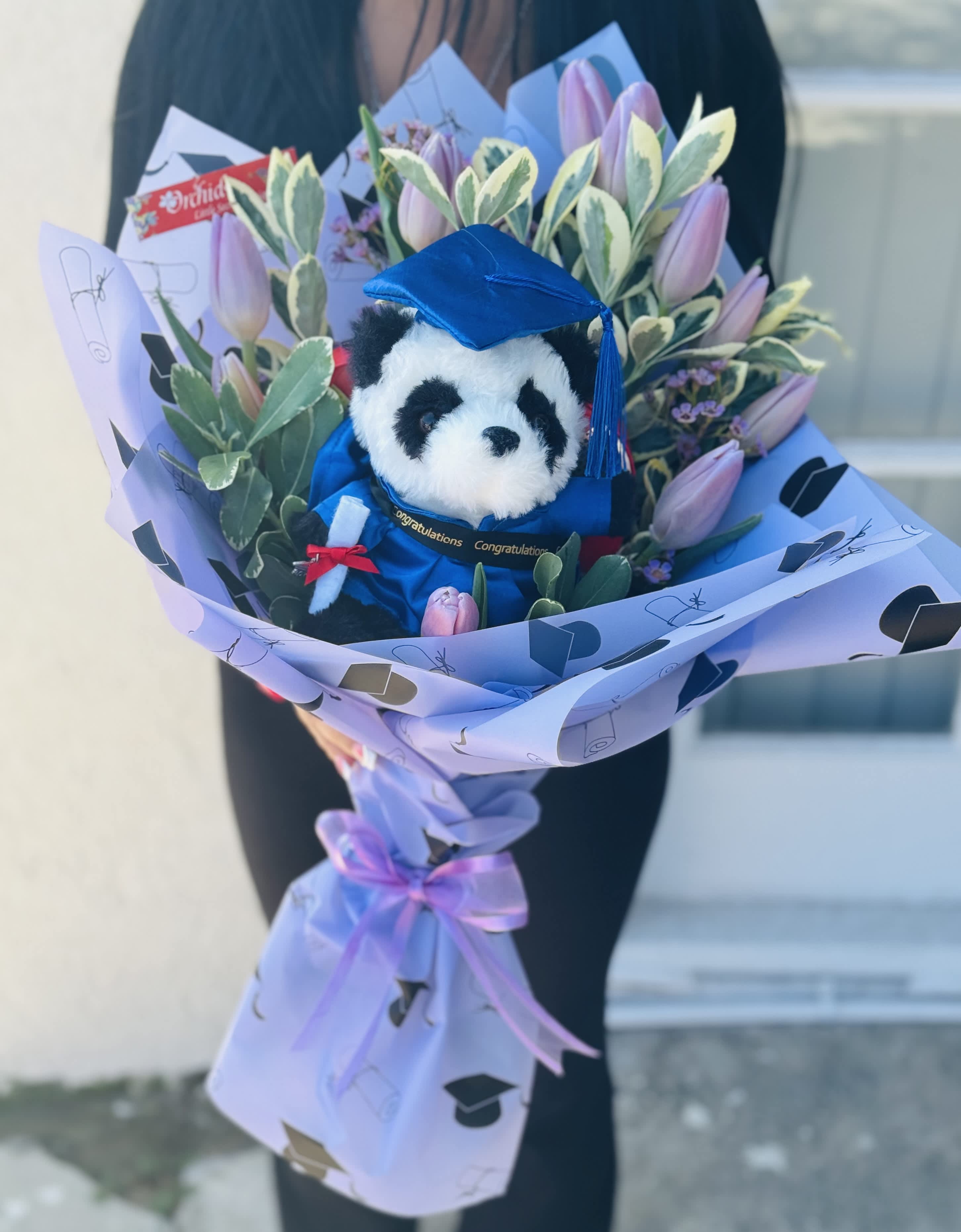 Bouquet with a plush panda in a blue graduation cap surrounded by pink tulips