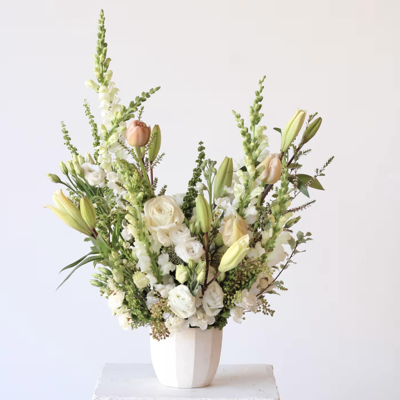 Tall white floral arrangement in a white vase with lilies, roses, and snapdragons