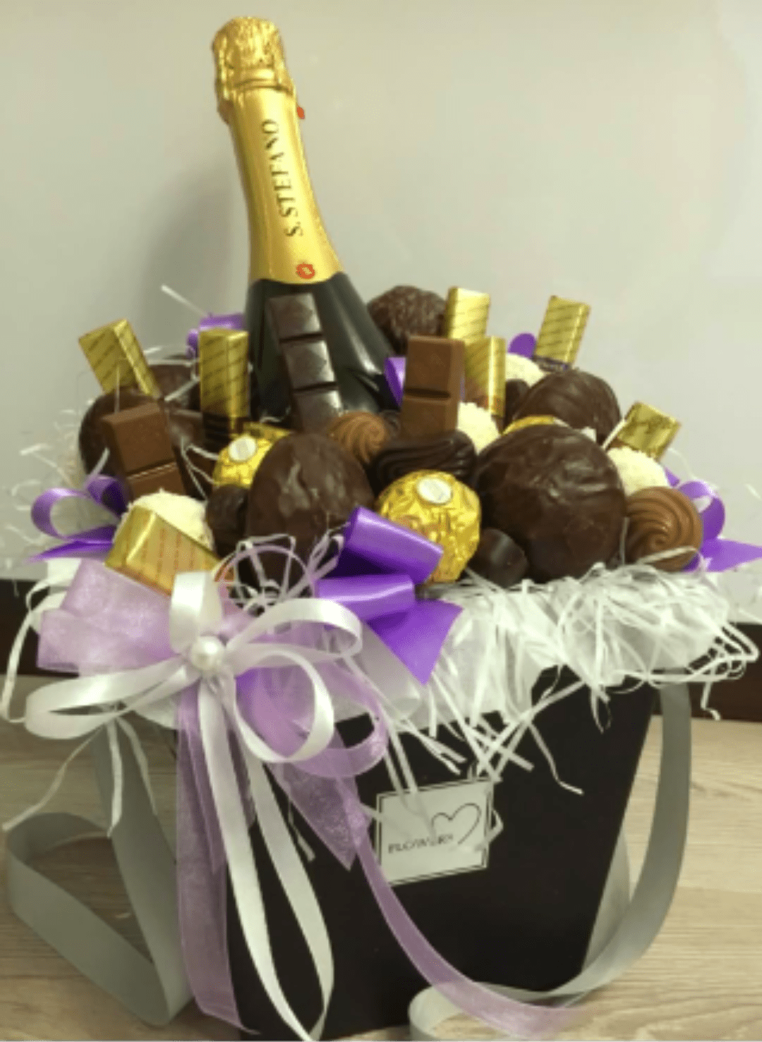 "Sweet Celebrations Gourmet Box" - The ultimate gift for a grand celebration. A chic black tote-style basket packed with a premium bottle of sparkling champagne, beautifully surrounded by an opulent assortment of gourmet chocolates. Featuring fine milk chocolate bars, gold-wrapped truffles, and decadent chocolate-covered confections nestled in crisp white shred.