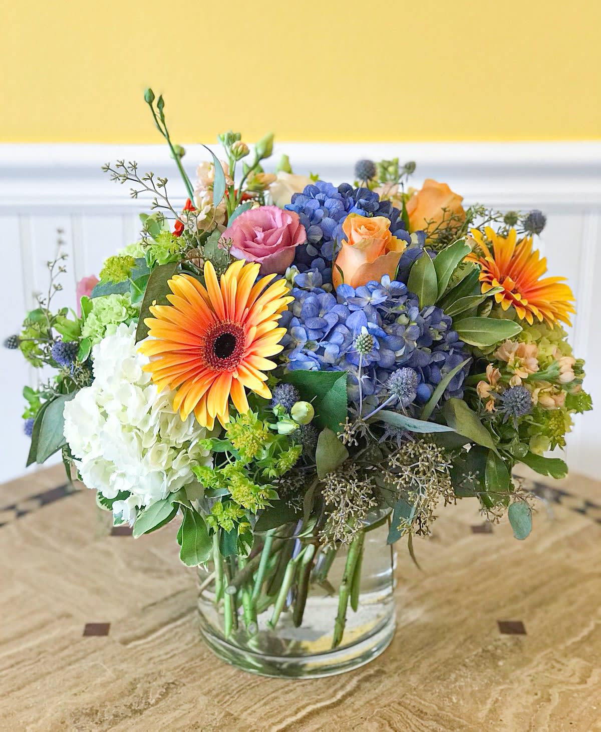 Mixed bouquet in a clear glass vase with blue hydrangeas, roses, and orange gerbera daisies