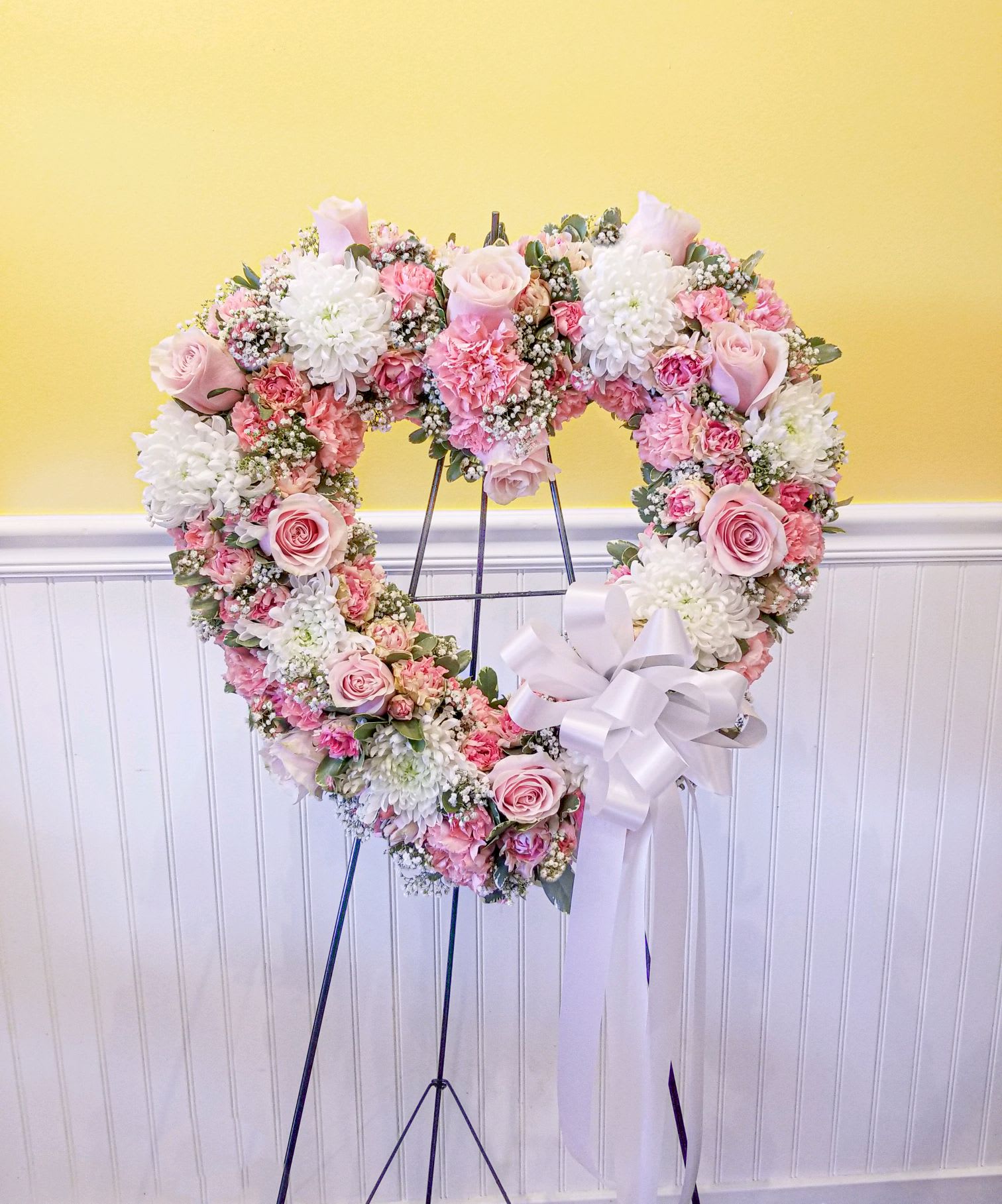 Heart-shaped floral wreath on an easel with pink and white flowers and a white ribbon