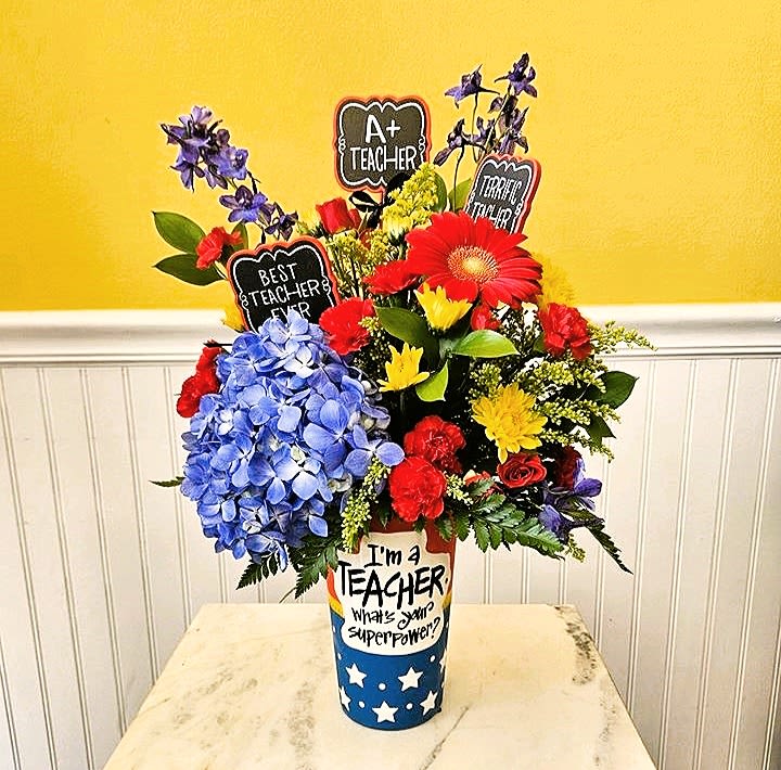 Bright mixed flower arrangement in a tall teacher-themed vase with chalkboard picks.