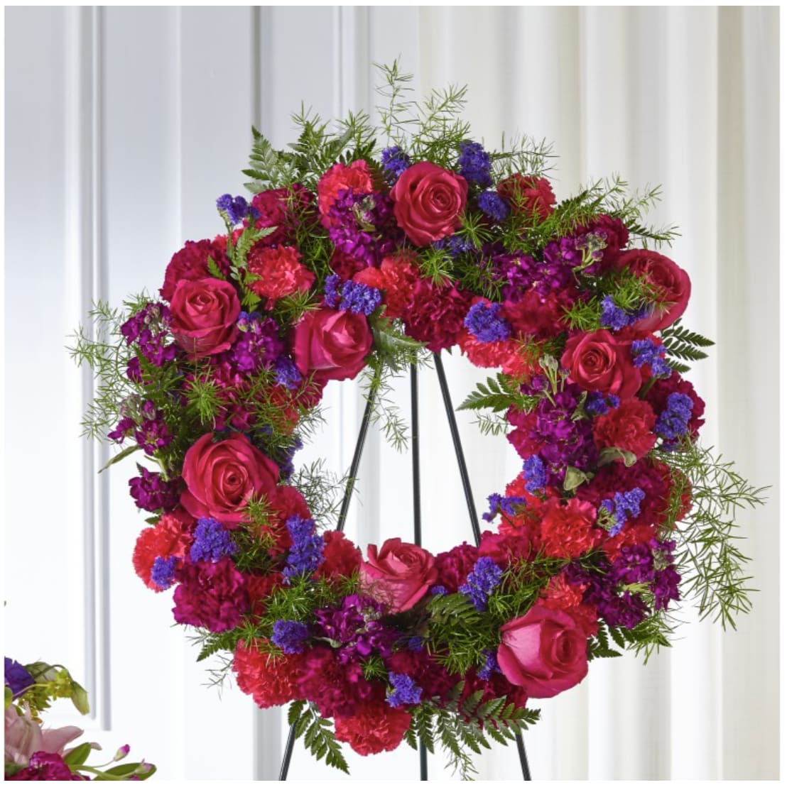 Circular floral wreath of pink roses, magenta carnations, and purple accents on a stand