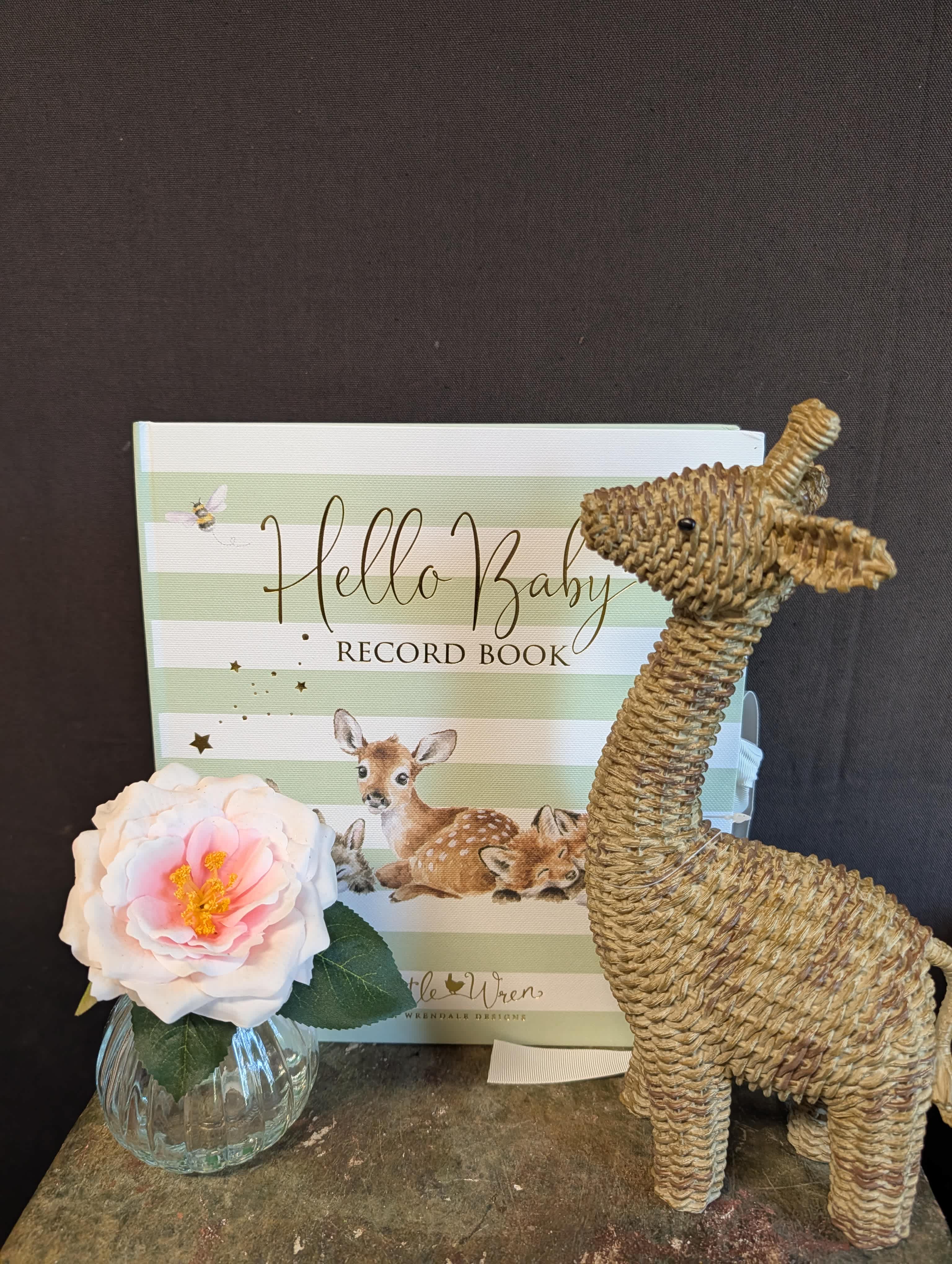 Single pink-and-white flower in a small glass vase beside a baby record book and woven deer figurine.