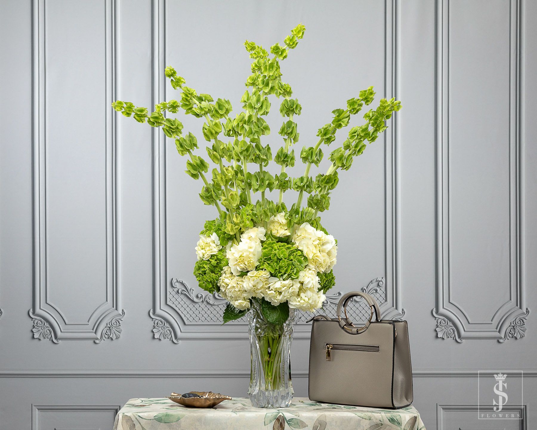 Tall green and white floral arrangement in a clear vase beside a beige handbag