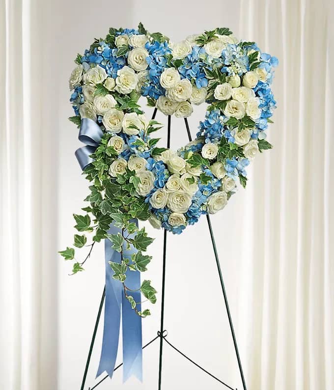 Heart-shaped floral wreath of white and blue flowers on a stand with a blue ribbon