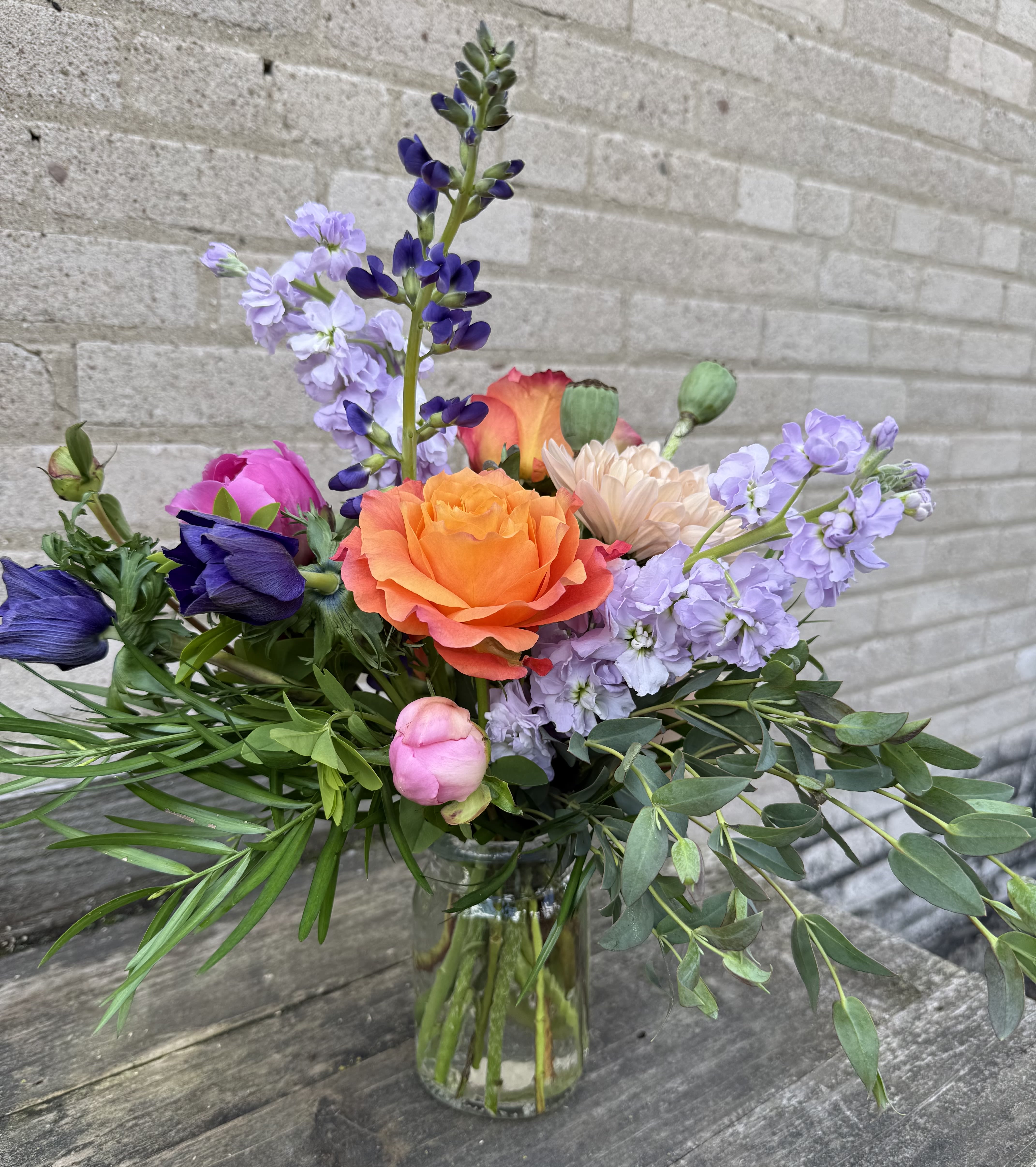 Summer Warmth - A vivid orange rose anchors this mixed-color flower arrangement, the perfect birthday bouquet or summer gift that feels both lively and romantic. Summer Warmth is ideal for celebrations from anniversaries and graduations to housewarmings, or as a cheerful get-well or thinking-of-you surprise that will make the person you're thinking of feel noticed and celebrated.  Pink roses and peach-toned blooms mingle with purple anemones, lavender and purple stock, and airy greenery in a loose, garden-style composition set in a clear glass vase. Tall purple and lavender spikes give height while lower flowers create a full, asymmetrical silhouette with a fresh, hand-tied appearance. The palette reads warm and festive-suitable for summer events like Labor Day or Fourth of July gatherings, yet equally at home for autumn holidays, Thanksgiving tabletops, or a bright touch during winter holidays.  Care is simple: keep stems trimmed and fresh water in the clear glass vase to prolong blooms for several days, and place out of direct heat. Available in one size shown here; order with a personalized note to mark birthdays, anniversaries, graduations, new babies, or just because moments when you want them to feel truly seen.