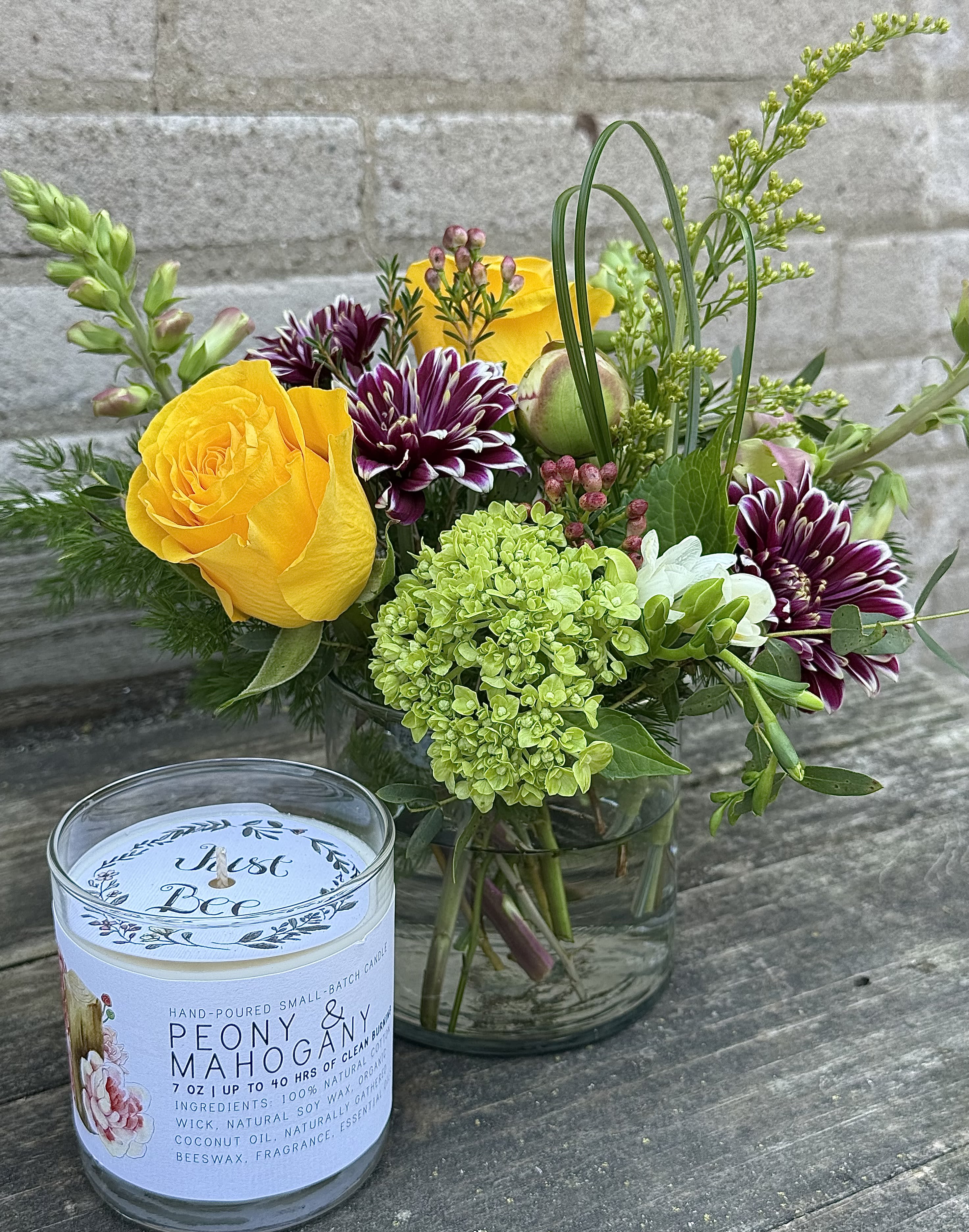 Flowers and a Scented Candle - Bright yellow roses and a rounded green hydrangea create an immediately cheerful focal point in this flower arrangement and scented candle gift set - perfect as a birthday bouquet, a thank-you gift, or a just-because surprise. The loose, garden-like design pairs sunlit roses with deep purple-and-white chrysanthemums and airy filler for a lively, slightly whimsical texture that will make the person you're thinking of feel genuinely celebrated. A white floral-scented candle in a clear glass jar adds a warm, finishing touch that extends the experience beyond the vase.  Arranged in a simple clear glass vase, the mix of roses, chrysanthemums, hydrangea, and other seasonal stems stands about a medium size and delivers layered color: sunny yellow, rich purple, crisp white, and fresh green. Tall accent stems give the composition an elegant, casual height while the rounded hydrangea balances the bouquet's silhouette. This set works well for occasions across the year - from Labor Day and autumn gatherings to housewarmings, anniversaries, get-well wishes, graduations, and holiday tables - and includes easy care: keep flowers in fresh water, out of direct heat, and enjoy the candle when you want a soft floral scent to linger in the room.