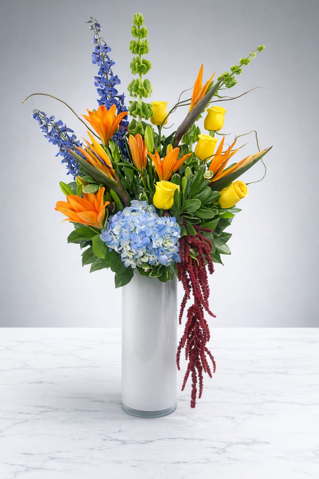 Tall mixed bouquet in a white vase with orange lilies, yellow roses, and blue flowers