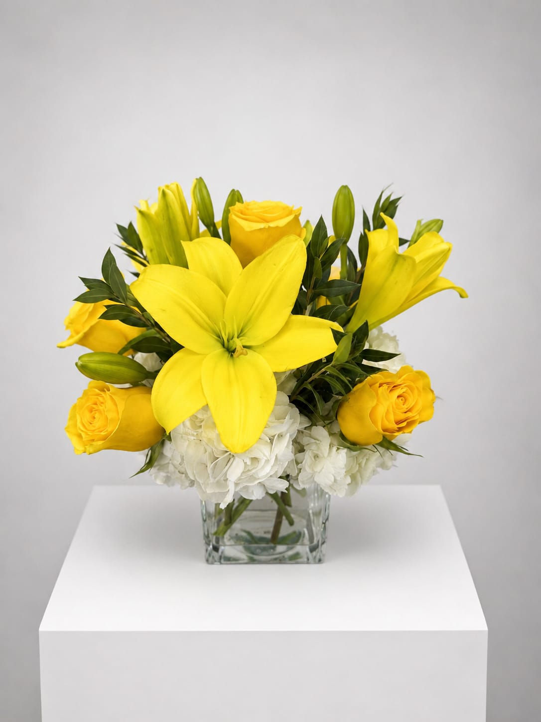 Yellow lilies and roses arranged in a clear square vase with white flowers below