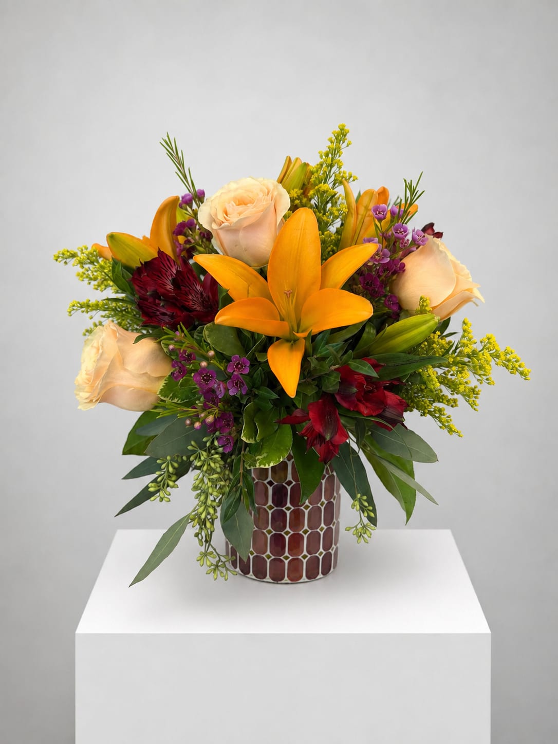 Mixed bouquet with orange lilies, cream roses, and purple accents in a patterned vase