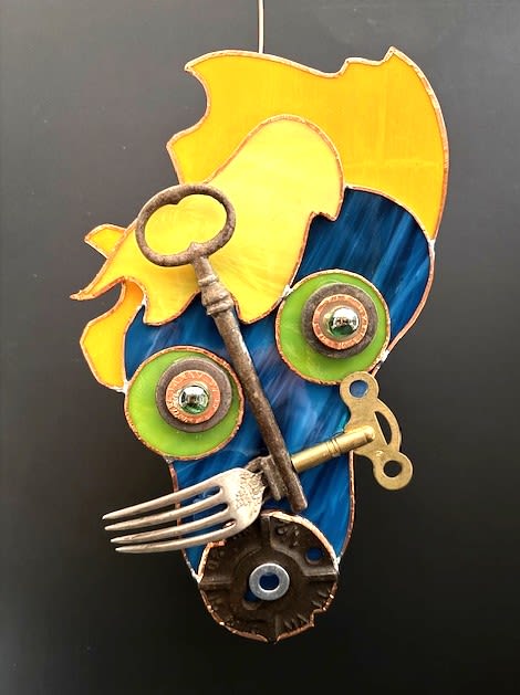 #A012 - "Totally Freaked Out" Mask - Bright yellow and deep blue stained glass sweep into a playful abstract face in this small wall art - a gift-ready piece perfect for birthdays or housewarmings. The design reads as a creative, mixed-color mask: two round green glass discs suggest eyes while layered metal washers and reflective caps add a gleam that catches the light. The overall effect is whimsical and lively, the kind of piece that makes the person you're thinking of smile every time they walk by.  Found-metal accents - a long skeleton key, vintage winding and clock keys, a decorative fork, metal gear and washers - are thoughtfully layered across the glass to create texture and a subtle steampunk edge. A stained glass plaque and hanging wire finish the piece so it's ready to display; the rustic metals contrast the bright glass for an eye-catching, eclectic look that suits studios, entryways, or creative living rooms.  This compact wall mask makes a distinctive, personal present for someone who collects unusual art or loves eclectic decor. It arrives ready to hang and is ideal for celebrating a birthday or marking a new home - a small but striking keepsake that will make the recipient feel truly seen.  Shipping available - select Delivery, then message us to ship.
