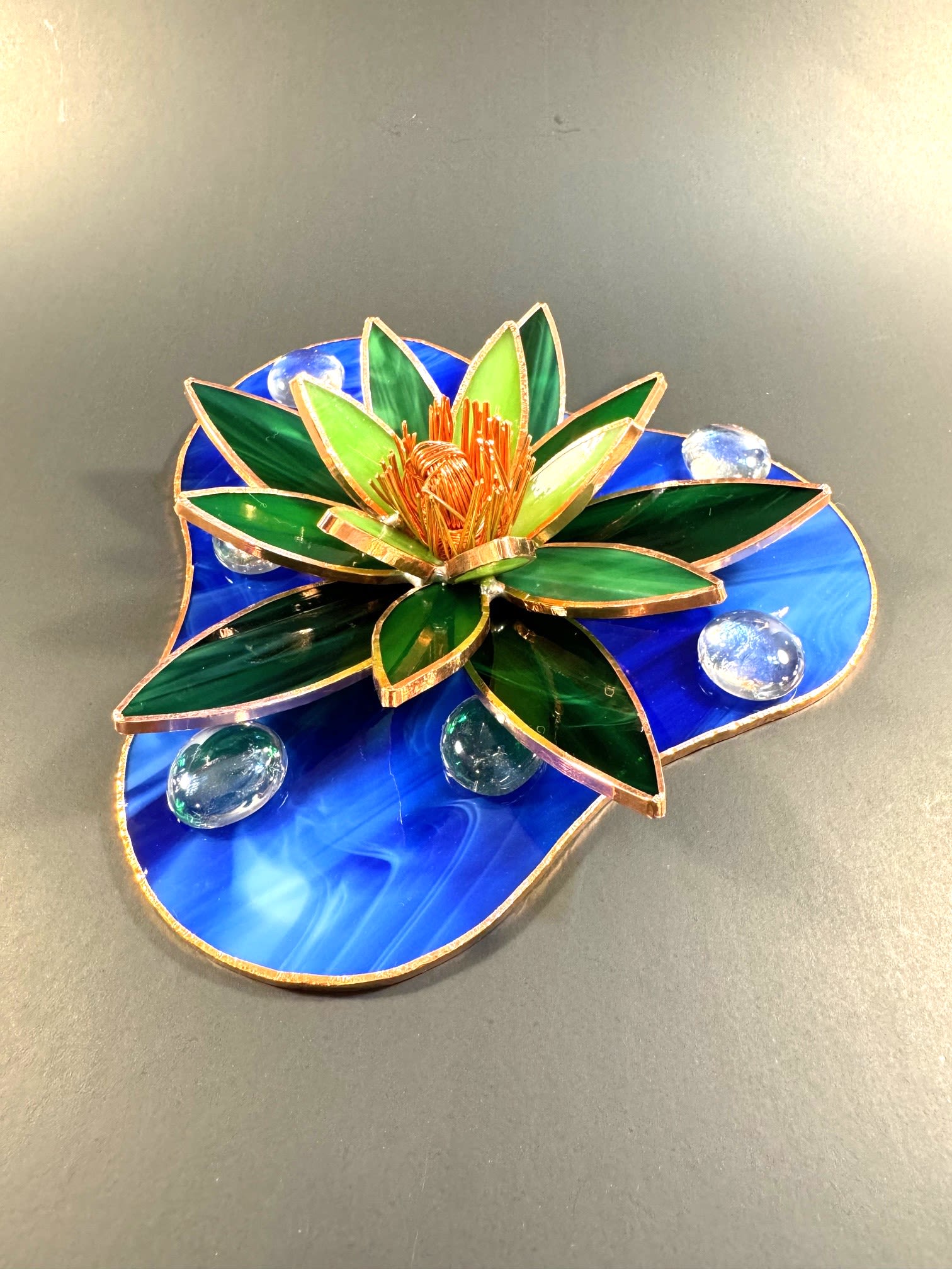 #A016 - Emerald Green &amp; Leaf Green Water Lily - A sculptural stained-glass water lily that becomes a calming focal point - perfect as a decorative accent or a thoughtful housewarming gift. This flower-style art piece reads as a green and blue lotus for home or office décor, ideal for someone who appreciates handcrafted glass art or wants a serene, reflective touch on a mantel, desk, or entry table. Layered petals of deep emerald and chartreuse glass fan out around a copper-toned, textured wire center, creating a dimensional, starburst effect that catches light. The flower rests on an irregular, marbled blue stained-glass base edged in metal to suggest gently moving water; clear glass pebbles and glass gems sit on the surface like sparkling droplets. Metal framework and copper wire detailing add warmth and structure, while the blue stained-glass slab anchors the piece visually.   Give this to the person you're celebrating when you want them to feel noticed and soothed - a lasting alternative to fresh flowers that fits birthdays, housewarmings, seasonal refreshes, or anniversary décor updates. Size and mounting details are shown on the product page; care is simple - wipe gently with a soft, dry cloth to preserve the glass and metal finishes.  Shipping available - select Delivery, then message us to ship.