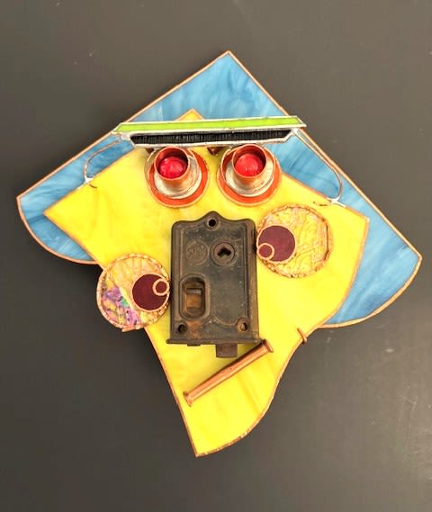 #A041 - "Glasses" Mask - A geometric, upcycled wall sculpture that reads like a playful portrait-this mask presents a stained glass "face" art piece ideal as a quirky gift or housewarming accent. As a decorative wall art or birthday present for someone who collects unusual objects, the stylized face immediately grabs attention: a yellow diamond-shaped panel edged in blue and copper-toned metal forms the head, while bold hardware elements read like facial features for an imaginative, conversation-starting display. The piece combines stained glass with repurposed metal details-two metal cylinders topped with red glass or plastic domes become wide, bright eyes; a vintage dark lock plate provides a nose-and-mouth anchor; iridescent round discs add cheek highlights; and a short copper pipe segment, thin metal rod, brush-tipped bar, small brush, decorative disks and a brush strip contribute texture and whimsy.   Colors include sunny yellow, cobalt blue, red accents and black metal tones, delivering a cheerful, tactile contrast. Perfect for the person who prefers art with personality, this wall sculpture suits living rooms, creative studios, or entryways where it can spark smiles and conversation. It arrives ready to hang and needs only a single nail or hook-no special care beyond occasional dusting-making it a low-maintenance, high-impact gift that will make the recipient feel seen and delighted by your thoughtful, offbeat choice.  Shipping available - select Delivery, then message us to ship.