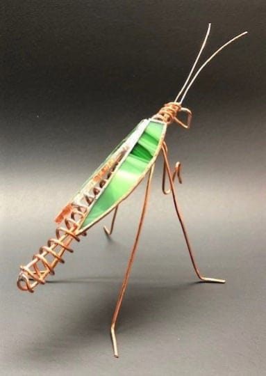 #A052 - Light Green Praying Mantis - A whimsical copper-wire insect sculpture shaped like a light green praying mantis makes an unexpected and delightful gift for someone who loves nature-inspired decor or a unique housewarming accent. This small decorative piece works well as a desk ornament, shelf accent, or tabletop focal point for birthdays, thank-you gifts, or casual celebrations-the kind of present that makes the person you're thinking of smile and sparks conversation.  Handcrafted from fine copper wire, the mantis features long, delicate legs, coiled tail detailing, and extended antennae that give it an airy, lifelike stance. The body is a triangular panel of variegated green stained glass set in a metal edge, with touches of orange and white reflected in the glass tones for extra visual interest. Lightweight and compact, it adds a playful, artistic touch without real botanicals and arrives ready to display among books, potted plants, or on a windowsill; no care required beyond occasional dusting.  Shipping available - select Delivery, then message us to ship.