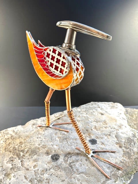 #A084 - Orange Door Handle Bird - A playful metal-and-glass bird that reads like a museum sculpture and a conversation starter - perfect as a housewarming gift or a cheerful birthday accent. This walking bird art piece pairs orange and red stained-glass panels with patterned metal sections to create a mosaic-like body, while a silvery antique car door handle suggests head and beak shapes for a whimsical, modern look. The warm color palette and quirky posture send a bright, lighthearted message that the person you're thinking of will notice.   The sculpture features long copper-tone legs with spiral-wrapped joints and splayed wire feet, giving it an animated, hand-crafted presence on a shelf, desk, or tabletop. Mixed-metal details and cutout patterns add texture and visual interest from every angle, so it reads as decorative home or office art that suits contemporary, industrial, or eclectic décor. An ideal pick for new-home celebrations, birthdays, or just-because gifting, this piece brings lasting color and personality without care concerns-place it where it can be admired and let its cheerful tones and lively form do the gifting for you.  Shipping available - select Delivery, then message us to ship.