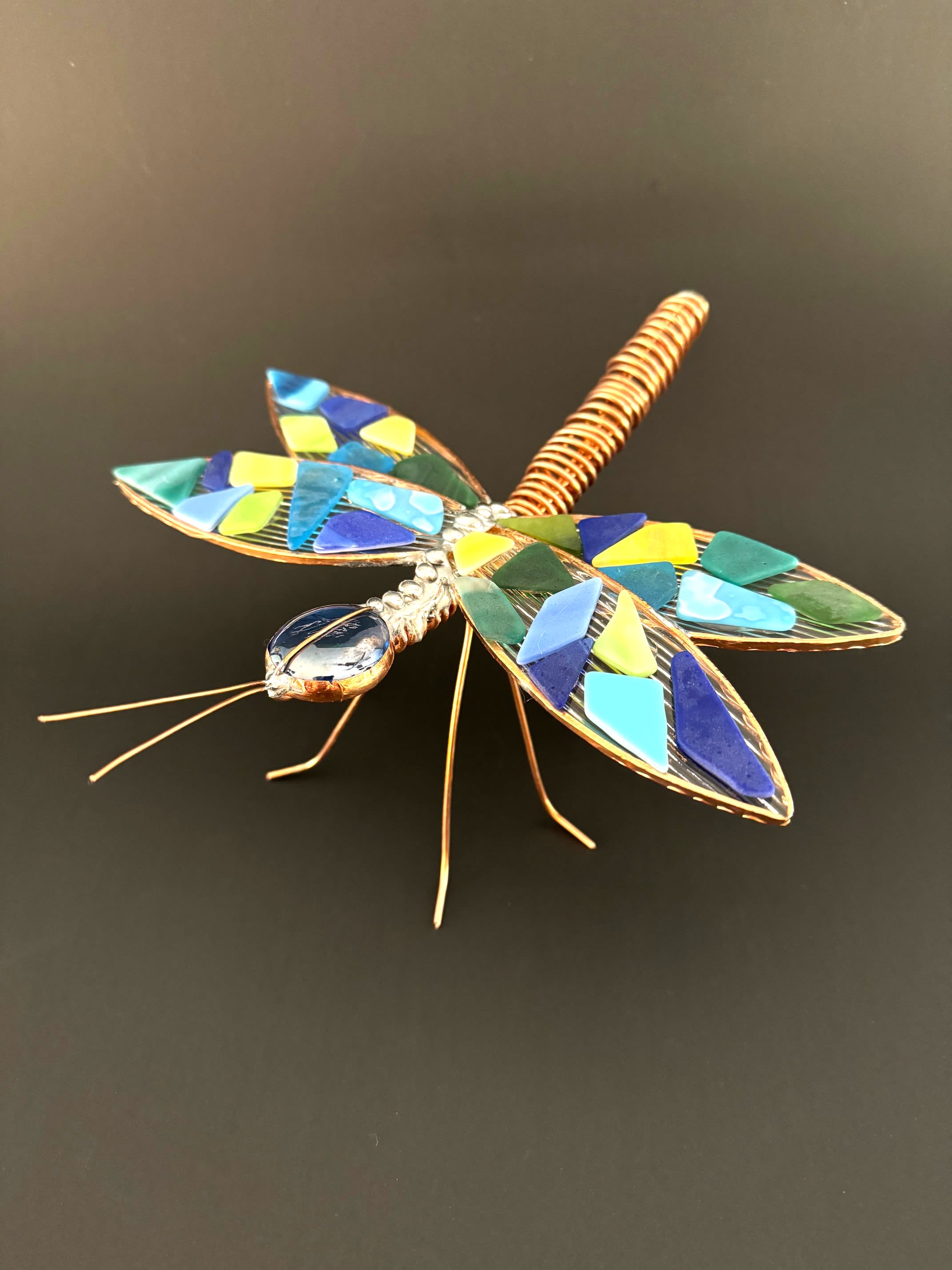 #A096 - "Water" Mosaic Dragonfly - A jewel-toned mosaic dragonfly adds a splash of playful artistry to any floral arrangement or gift for a nature lover. This decorative accent-perfect for springtime celebrations, housewarmings, or as a small keepsake - puts the sculpture and occasion (home, garden, seasonal gift) front and center so the person you're thinking of feels seen and delighted. Handcrafted metalwork forms a coiled copper-toned body with fine wire legs and antennae, topped by a rounded translucent blue glass head that catches light.   The wings are adorned with irregular glass mosaic pieces in blues, teals, greens and bright yellow, producing a jewel-like, geometric pattern that contrasts beautifully with a matte dark background. The piece reads as both artisan décor and a cheerful floral topper; it works equally well tucked into a bouquet or displayed alone on a shelf. Give this dragonfly as a thoughtful accent for birthdays, anniversaries, or to brighten a get-well or thinking-of-you bouquet-the vivid colors and handmade feel convey warmth and cheer. Sized to complement most arrangements, it requires no care beyond gentle dusting and will keep brightening the recipient's space for years to come.  Shipping available - select Delivery, then message us to ship.