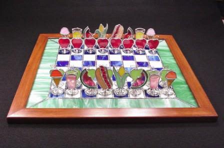 #A101 - Summer Picnic Chess Set - A sunny burst of color and whimsy, this one-of-a-kind stained-glass Summer Picnic chess set is a playful take on decorative gifts for housewarmings or summer celebrations. The decorative chess board is framed in warm wood with alternating deep blue and white glass squares, ringed by a marbled green border that reads like a lawn around a picnic blanket. The recipient will see it and smile: it's the kind of artful accent that makes a living room, sunroom, or entry table feel immediately more personal. Each "piece" is a tiny stained-glass sculpture-cheerful red and green apple motifs, colorful cocktail glass shapes, fruit slices, and cone-like treats - mounted on clear round bases so they sit like little sculptures across the board. The mix of blue, green, red, and white glass gives strong visual contrast and texture; the wooden frame adds warmth and makes the set ready to display.   It's intended as a decorative conversation piece rather than a competitive game set, it's perfect for gifting to someone who loves artisanal home décor. Give this to the person who delights in quirky, handmade accents-ideal for birthdays, housewarmings, or a cheerful "thinking of you" surprise. Treat it as a display object: wipe the glass gently with a soft, dry cloth and avoid abrasive cleaners to preserve the finish. Orders ship ready to place on a mantel, coffee table, or shelf where it can catch the light and start conversations.  Shipping available - select Delivery, then message us to ship.