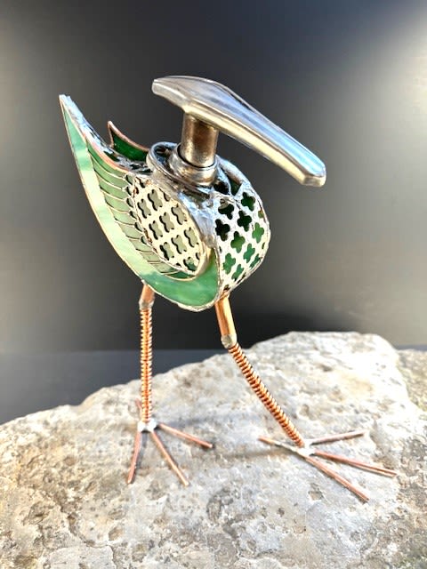 #A114 - Green Door Handle Bird - A whimsical mixed metal bird that reads like modern folk art.  This standing sculpture features a sculptural bird accent perfect for gifting on a birthday, housewarming, or as a unique thank you. The piece's green stained glass wings and patterned metal body catch light and add a joyful pop of color; placed on a shelf or tabletop it instantly becomes the kind of object that makes the person you're thinking of smile. Crafted from layered metal panels, translucent green stained glass, and polished silver tone details, the bird stands mid stride with long coiled copper legs and simple wire feet.   Stained glass panels and mosaic like cutouts give depth and shimmer, while a smooth metal handle repurposed as the beak and complementary stained glass accents and black details create a modern, artistic contrast. The sculpture's compact scale makes it easy to display on a desk, mantel, or entry table. This playful, keepsake worthy accent is ideal for someone who appreciates handcrafted, artful decor-a meaningful way to mark a special moment or to give a memorable housewarming gift. Because it's all solid materials, no special care is needed beyond dusting; choose it when you want a lasting, decorative present that will be enjoyed for years.  Shipping available - select Delivery, then message us to ship.