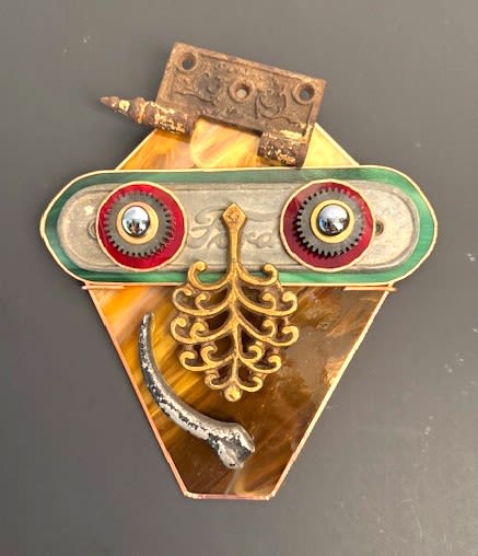 #A119 - "Thief!" Mask - A whimsical steampunk-inspired wall sculpture that reads like a quirky mask - perfect as an offbeat gift for a birthday or housewarming. This small assemblage art piece uses a marbled amber glass plaque and amber-colored backing plate as the warm, tapered base, while rusted hinges, a horizontal metal brow, red-rimmed gears and domed screws form a playful pair of eyes and a pronounced forehead. The overall effect is bold and tactile, the kind of object that will make the person you're thinking of smile every time they pass it.   Mixed-metal textures in orange, red, yellow and green tones give this piece a rich, vintage-industrial look: antique filigree elements hang centrally like a nose or mustache, shiny metal ball bearings sit at the gear centers, and an antique car door handle suggest an expressive mouth. Finished with rusted hardware, decorative filigree and a curved metal rod, it arrives ready to hang as a small wall-mounted sculpture. Give it to someone who delights in found-object art-this mask-like accent will add character to an entryway, studio, or eclectic living space.  Shipping available - select Delivery, then message us to ship.