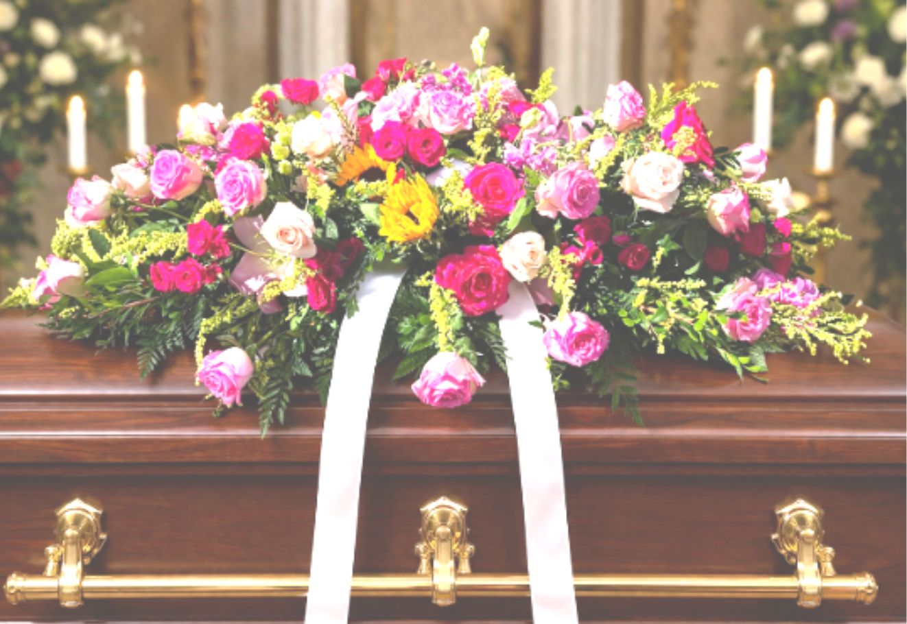 Bright pink and cream rose casket spray with white ribbon on a wooden casket.