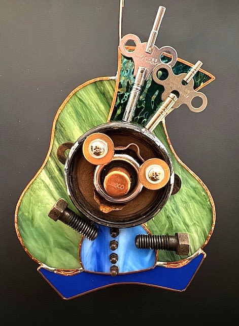 #A097 - "Joyful Little Guy" Mask - Swirled green stained glass forms the cheerful torso of this small mixed-media wall-style art piece, while cobalt blue glass suggests a jacket or grounded base-perfect as a birthday gift or a playful housewarming token. The circular face is built from an assortment of washers and round metal pieces, with bolts, screws, and wind-up keys arranged as arms, buttons, and whimsical antennae; the overall effect is a joyful, steampunk-inspired character that will make the person you're thinking of smile.   The contrast of smooth, translucent glass and textured recycled metal gives this decorative accent rich tactile appeal: green, blue, yellow, orange and black highlights catch the light from different angles, and a metal cup and stained-glass plaque complete the composition. Sized as a small tabletop piece, it's ideal for desks, shelves, or entryway tables-an unconventional, creative keepsake that says "you stood out to me" without saying a word. Care is simple: dust gently with a soft cloth and avoid prolonged exposure to direct sunlight to preserve the glass colors.  Shipping available - select Delivery, then message us to ship.