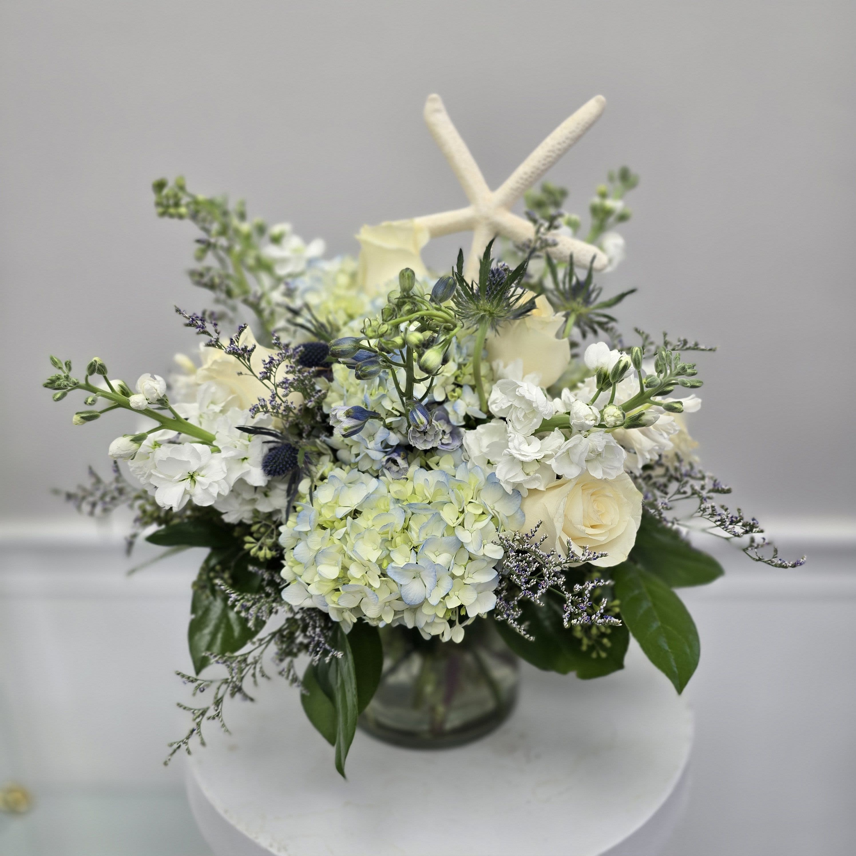 Ocean Breeze - This Wonderful Bouquet features Blue and white flowers including Hydrangeas, Delphinium, Thistle and a star fish.     *Container and Flowers may differ slightly from what is shown in the picture depending on availability and season