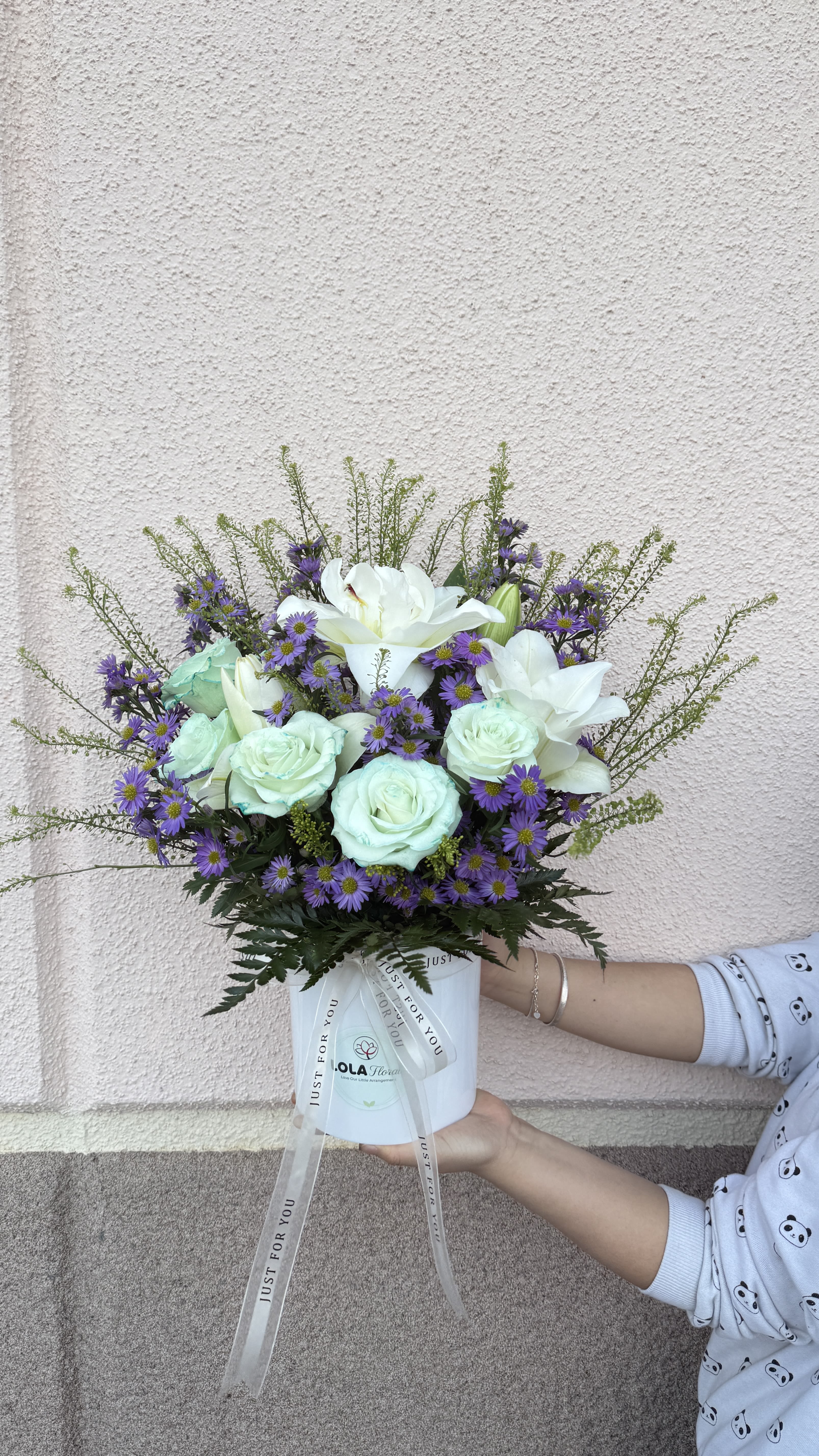 Box bouquet 01 - Soft white lilies and pale mint roses spill from a crisp white hatbox in a compact, garden-style arrangement-an elegant flower arrangement perfect for birthdays, thank-you gifts, or spring celebrations. The cool white and lavender tones, lifted by small purple aster-like blooms and touches of pink-green foliage, give the recipient a softly romantic, fresh impression that reads thoughtful without being overdone.  Arranged low in a white hatbox and tied with a long satin ribbon, this design balances airy filler with lush focal blooms for a full, textured look that feels both polished and effortless. The mix of lilies, roses, and small daisy-like asters creates gentle contrasts of shape and scent; the hatbox presentation makes it easy to place and enjoy right away. Ideal for the person you want to make feel seen-a sweet, versatile gift that arrives ready to display.