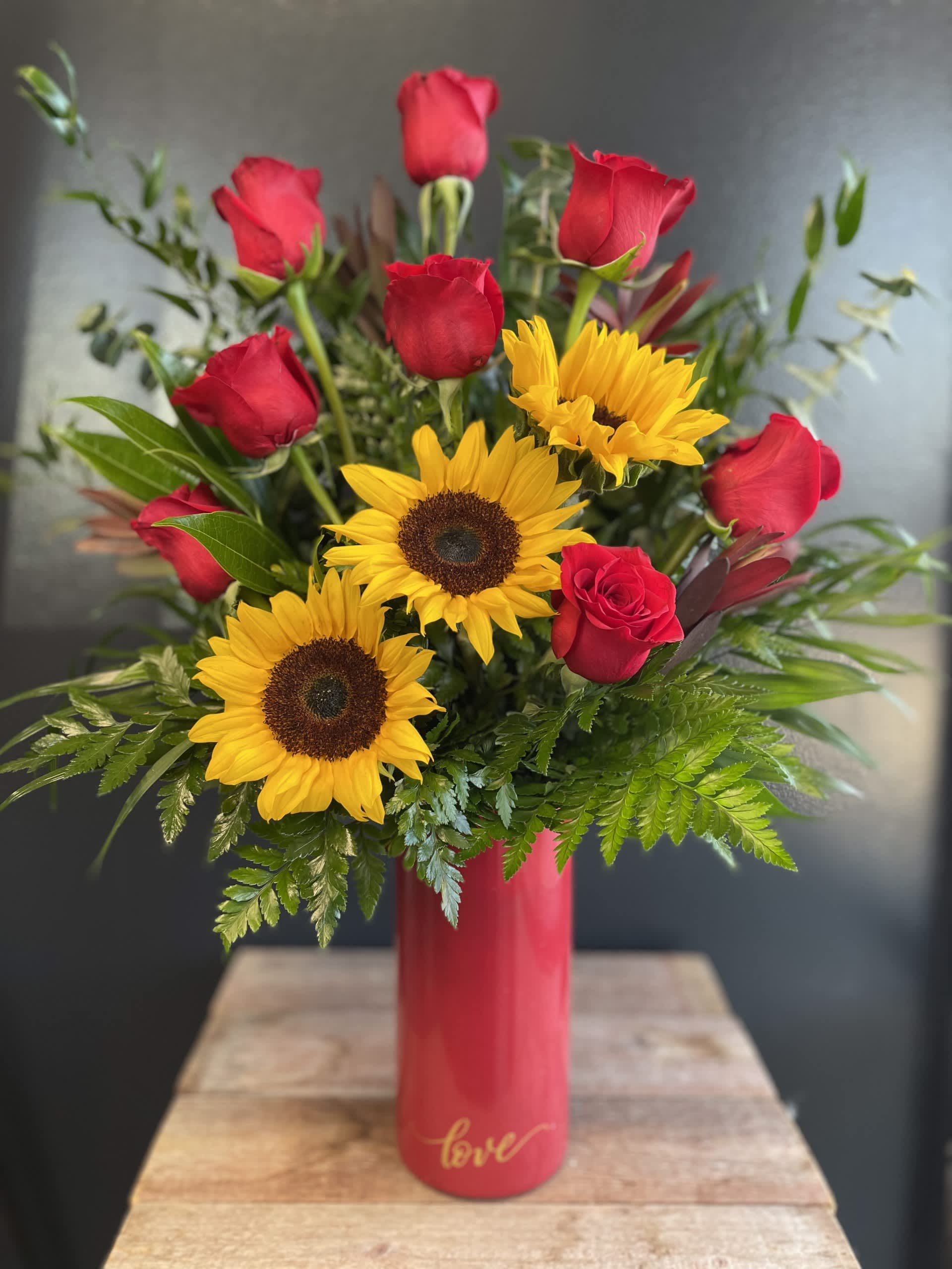 Sunflowers and Roses  - Experience the perfect harmony of vibrant sunflowers and passionate red roses in our Sunflower and Red Rose Floral Arrangement. This striking combination blends the cheerful warmth of golden sunflowers with the timeless elegance of deep red roses, creating a stunning display that symbolizes both joy and love. Red Vase is currently unavailable, substituted with a clear vase at this time. 
