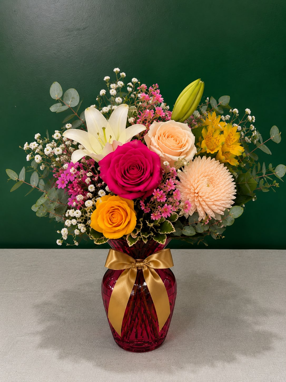 Mixed bouquet of roses, lilies, and daisies in a red vase with a gold ribbon