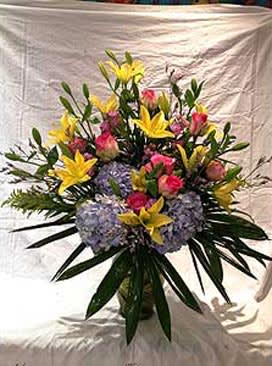 Tall arrangement of yellow lilies, pink roses, and pale lavender hydrangeas in a glass vase