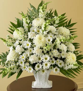 Tall white sympathy arrangement of daisies, mums, and snapdragons in a white urn-style vase