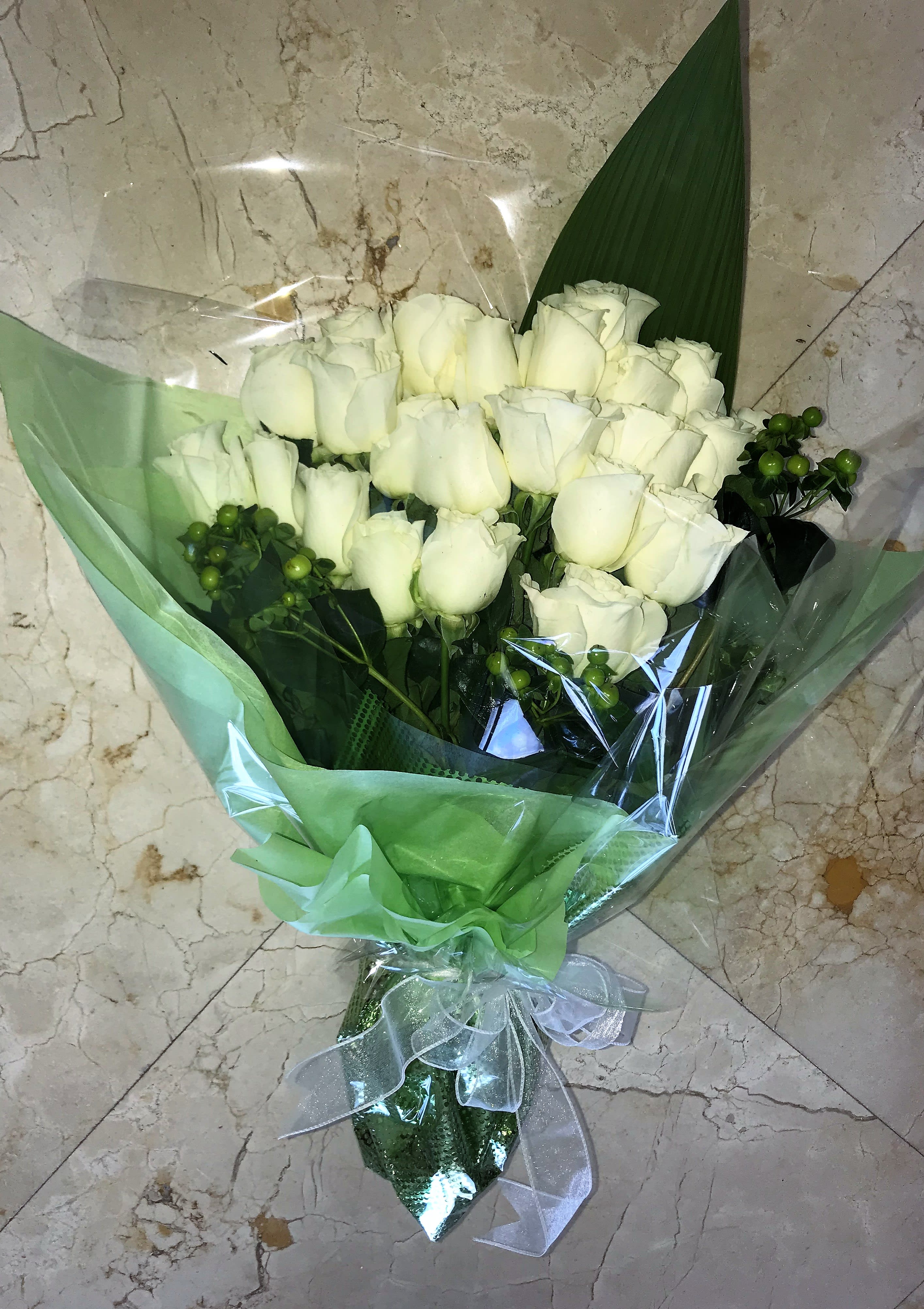 Bouquet of white roses with green berry accents wrapped in green and clear cellophane.