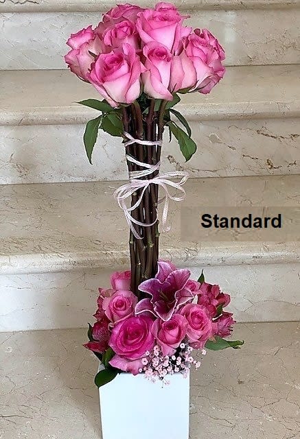 Tall arrangement of pink roses and lilies in a white square vase with bundled stems tied with ribbon