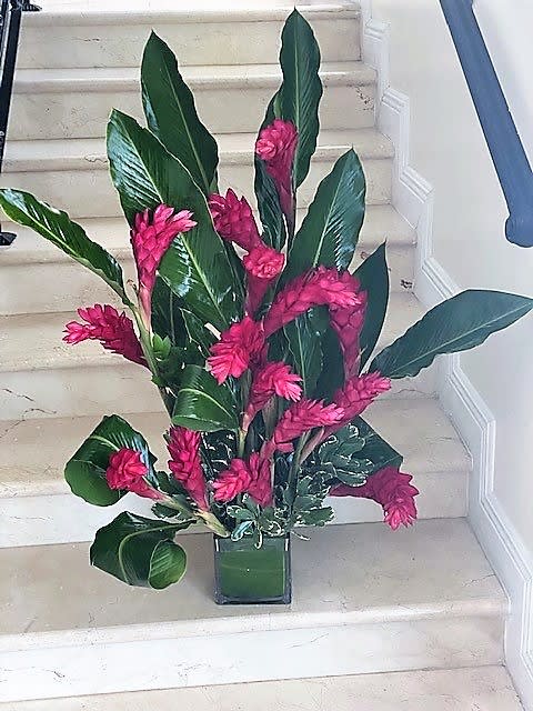 Tall tropical arrangement with bright pink flowers and large green leaves in a square glass vase