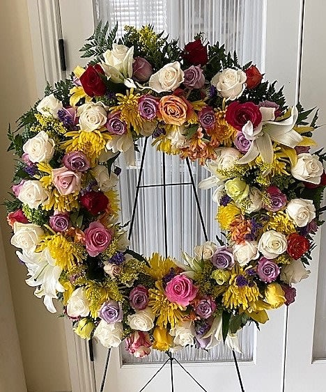 Large circular standing wreath of mixed roses, lilies, and yellow mums on a metal easel stand