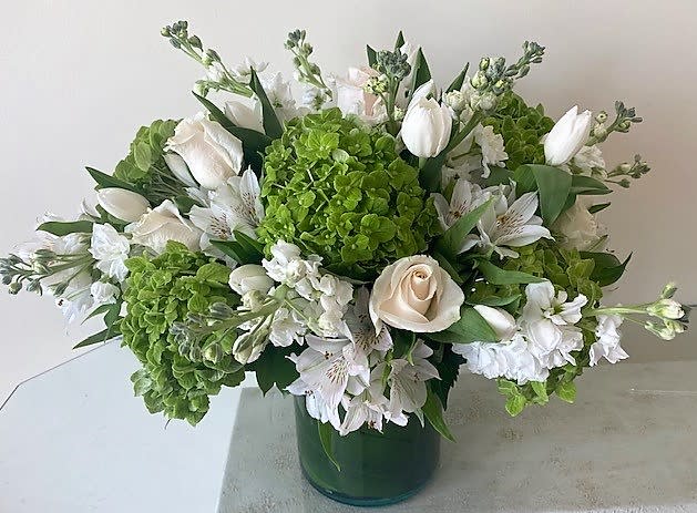 Low arrangement of white roses, tulips, and green hydrangeas in a green glass cylinder vase